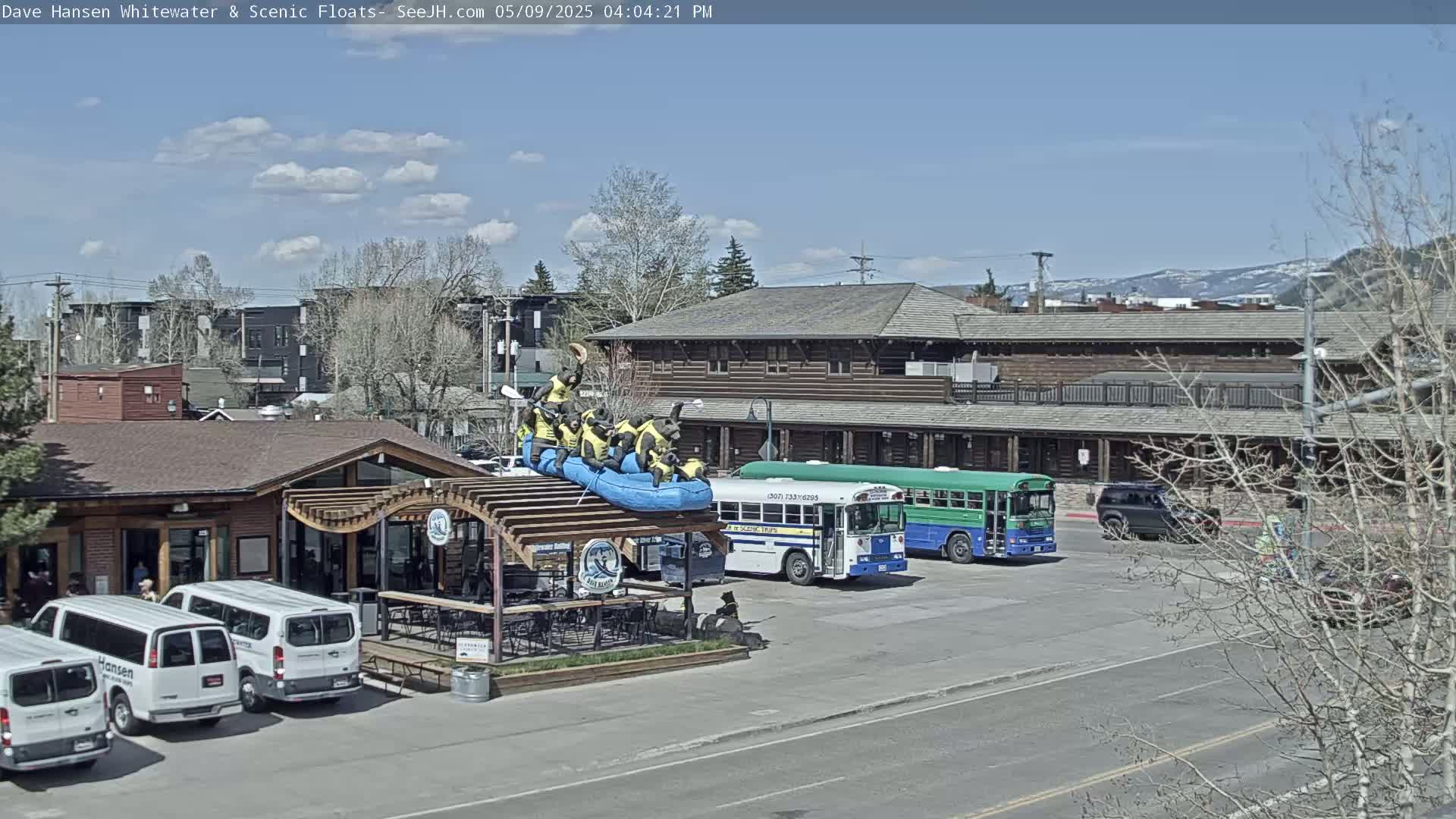 A partly cloudy blue sky is above a street scene with a building, several vehicles including buses and vans, and a raft with figures on top of a pergola.