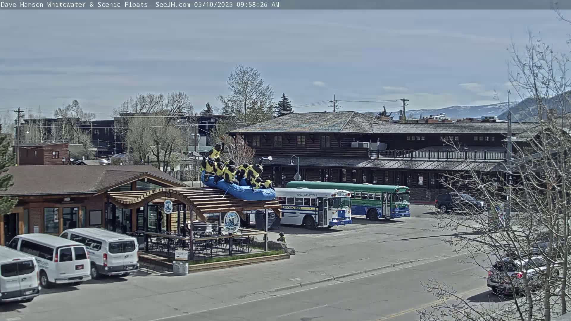 A sunny day reveals a town scene with a restaurant patio, several parked vans, two school buses, and a raft with figures atop a building.