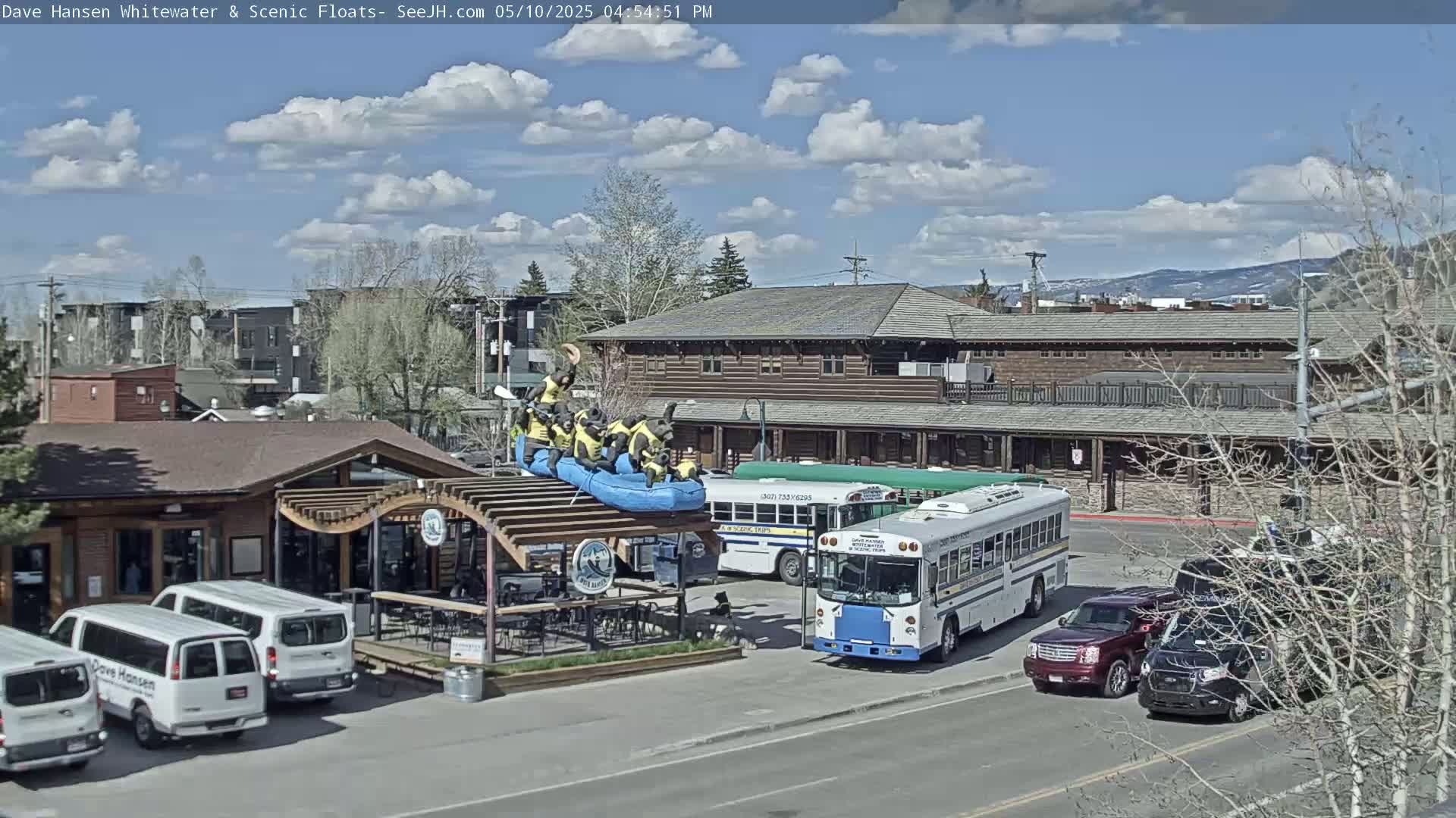 A partly cloudy day shows a building with several vehicles parked in front, including vans, buses, and SUVs, plus a raft with figures on top of a pergola structure.