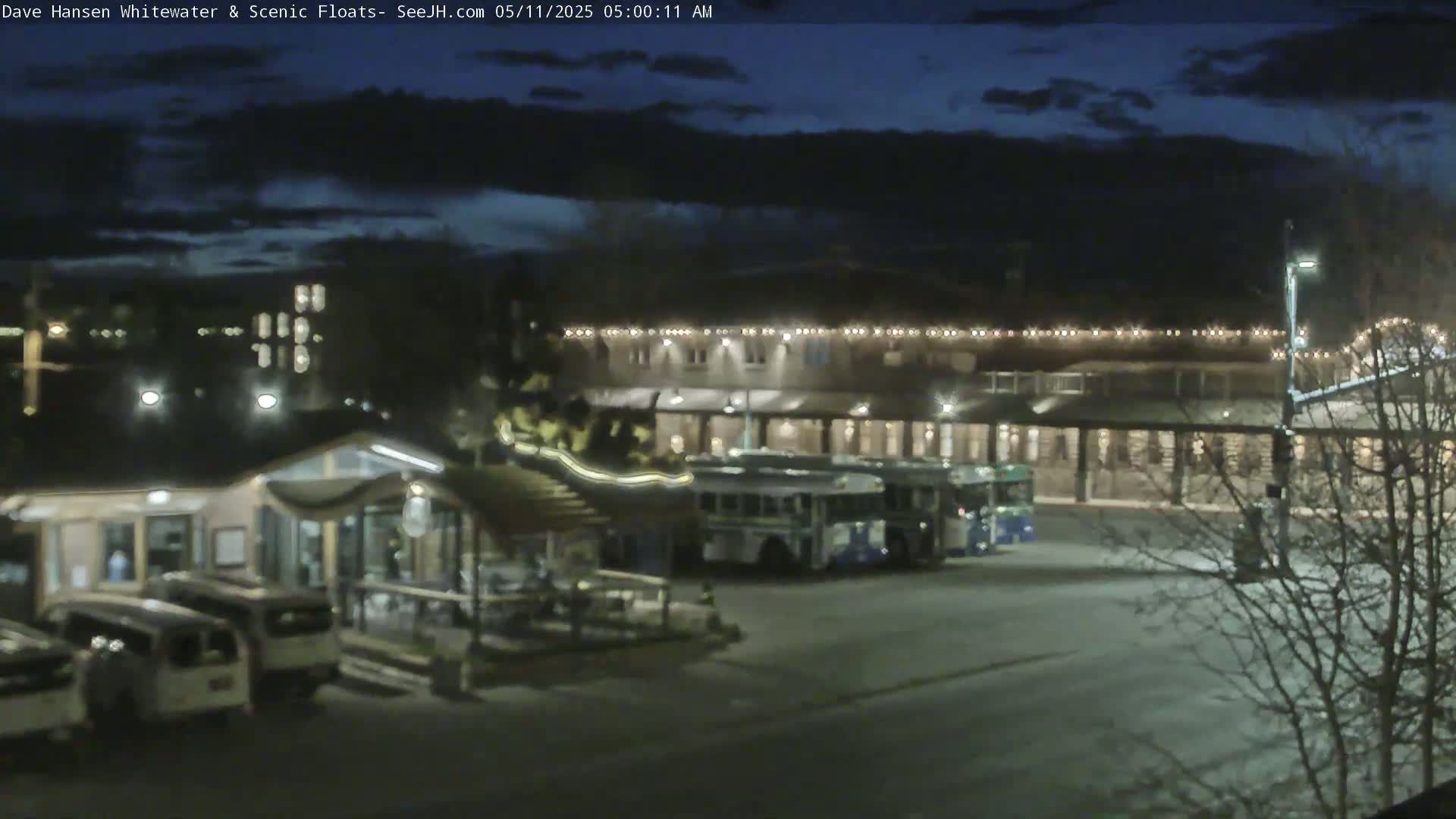 Under a dark, cloudy night sky, several buses are parked near a building with string lights, alongside a smaller structure and several vans.