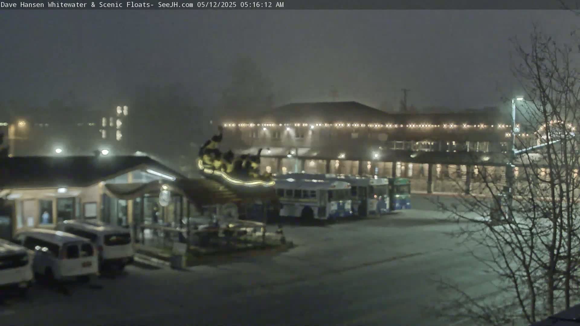 A nighttime, foggy scene shows several vehicles parked near a building with string lights, and a long bus parked in front of it.