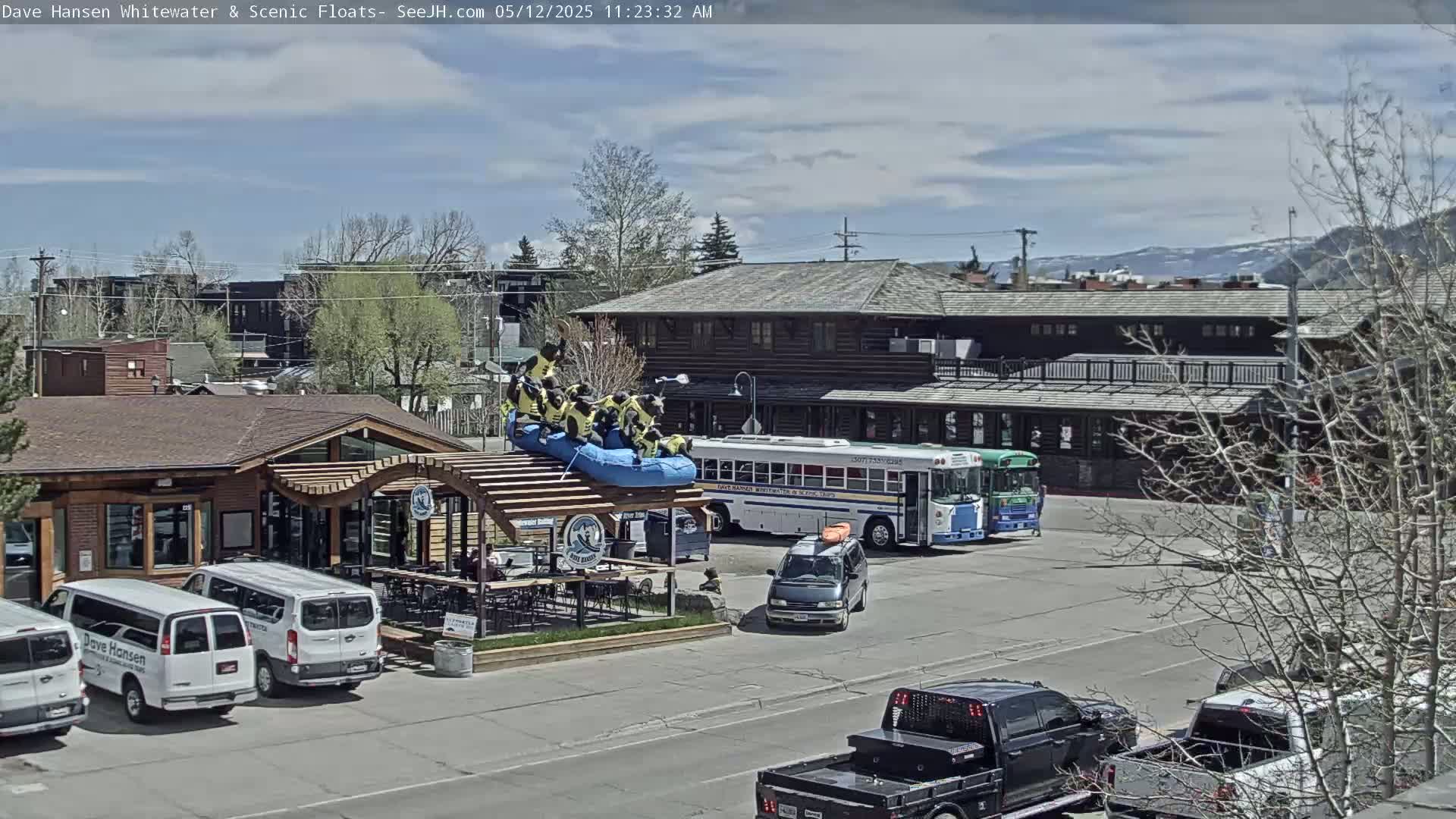 A group of people in yellow life vests are sitting on a blue raft on the roof of a wooden structure; several vehicles are parked nearby under a partly cloudy sky.