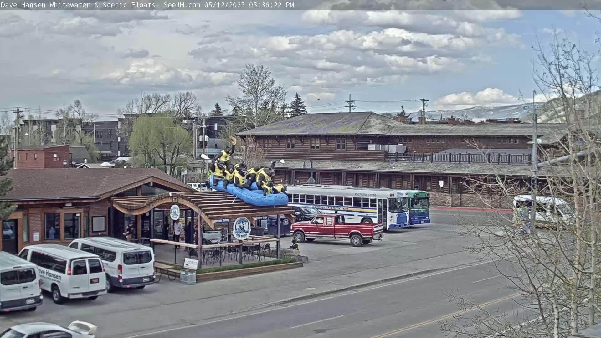 A partially cloudy day shows a raft with people in yellow life vests on a building's roof, near parked vans, a red pickup truck, and a bus in a parking lot.