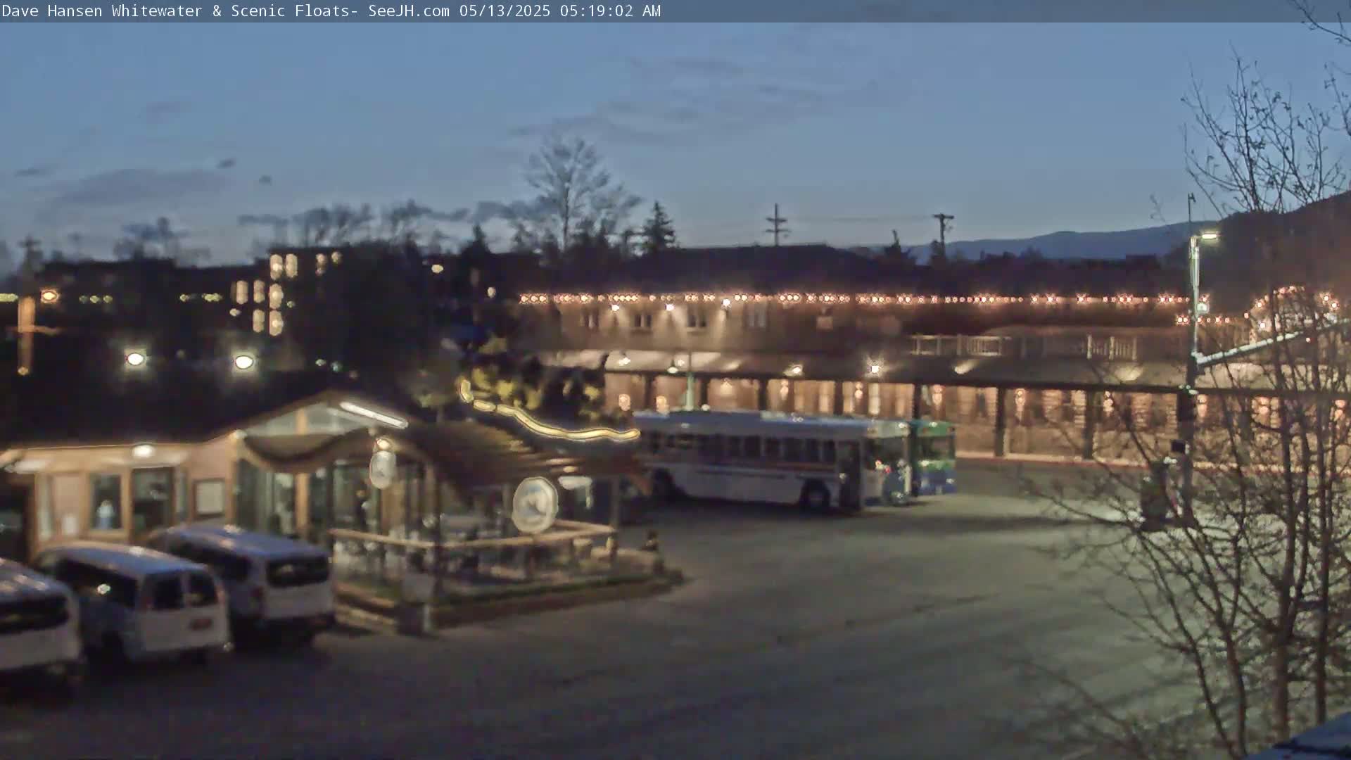 A dimly lit parking lot at twilight, with several cars and a bus parked near a building decorated with string lights.