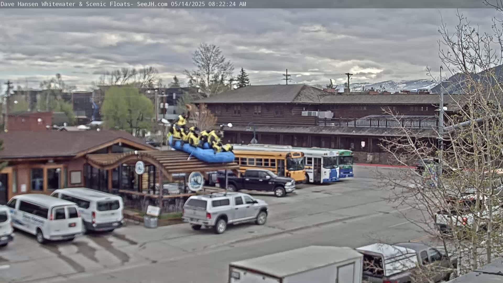 A group of people in yellow life vests sits atop a blue raft on a covered structure near a parking lot with several vans, trucks, and buses, under an overcast sky with mountains in the distance.