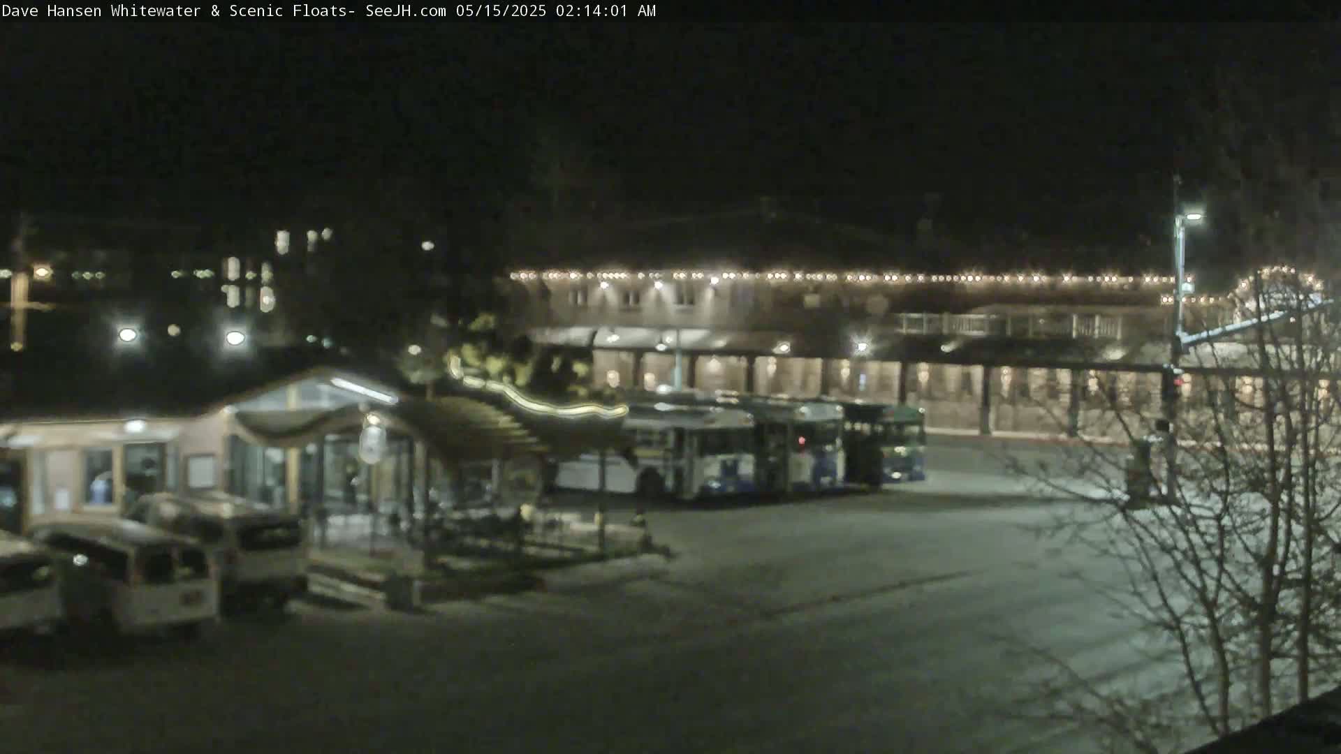 At night, several buses are parked in a lot next to a building with lights strung across its facade, and some cars are parked near a small building.