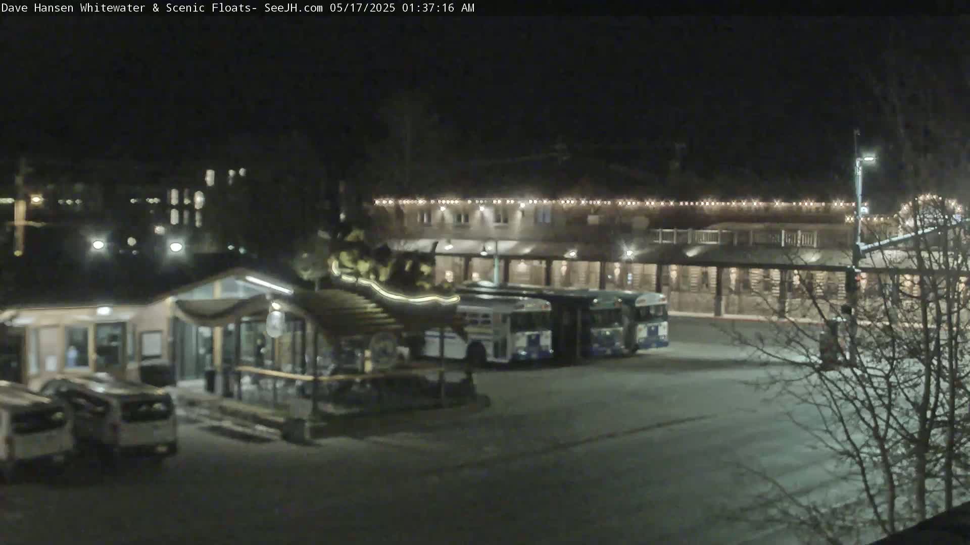 At night, several buses are parked in a lot next to a building with lights outlining its roofline.