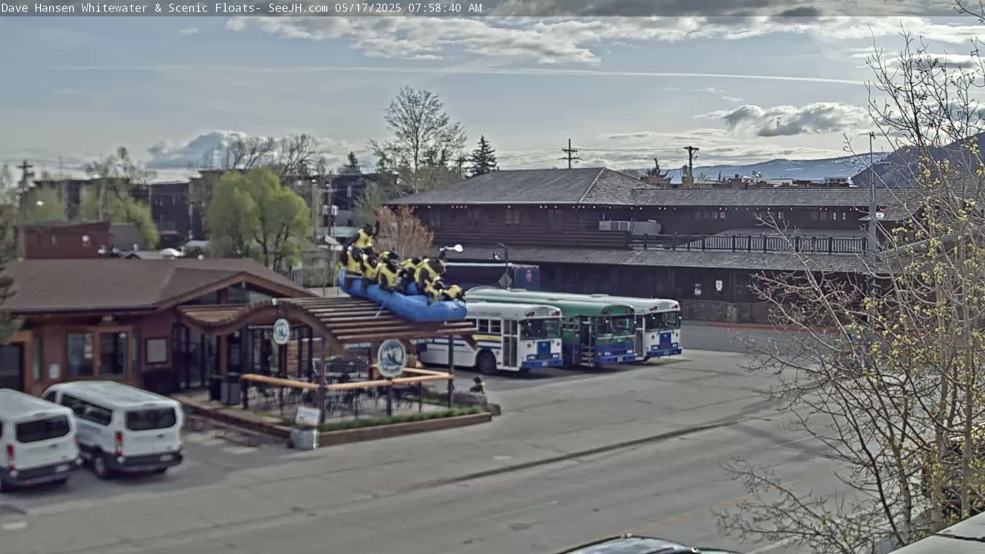 A partly cloudy day reveals a scene of a building with a patio, two vans parked in front, three buses parked nearby, and a raft with people on top situated on a structure above the patio.