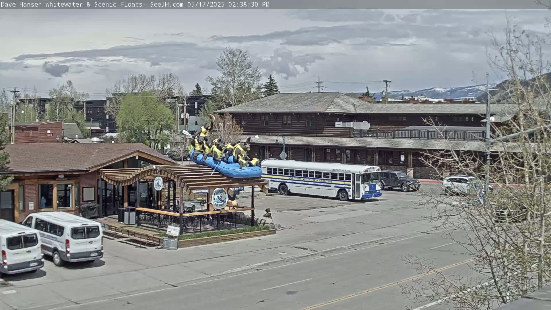 A partly cloudy day shows a building with a patio where a raft with bear figures sits atop a pergola, a school bus and several cars parked in a lot, and additional buildings in the background.