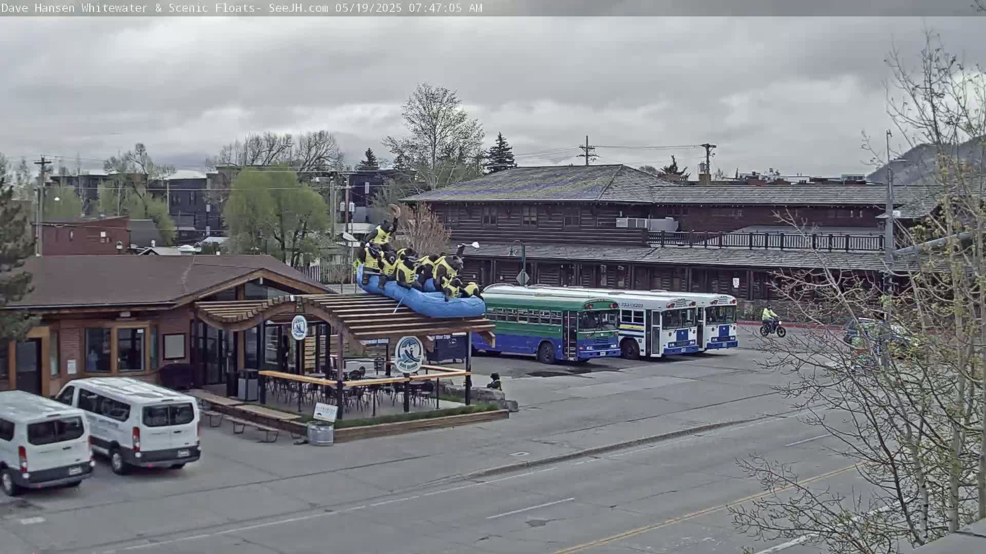 A raft with people in it sits on the roof of a building near two buses and some vans under an overcast sky.