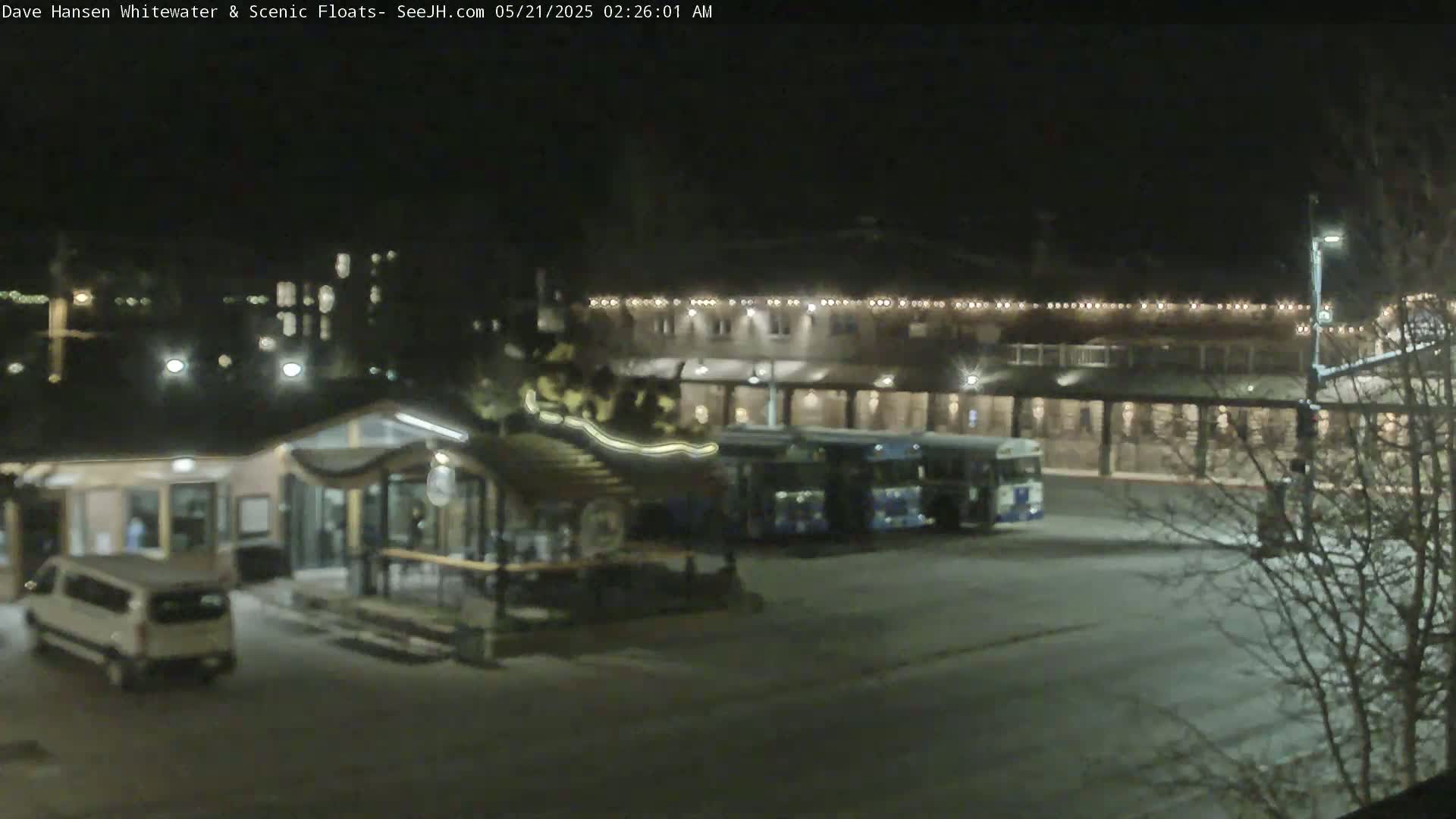 A nighttime, snow-covered parking lot with a building, a small kiosk, a van, and two buses.