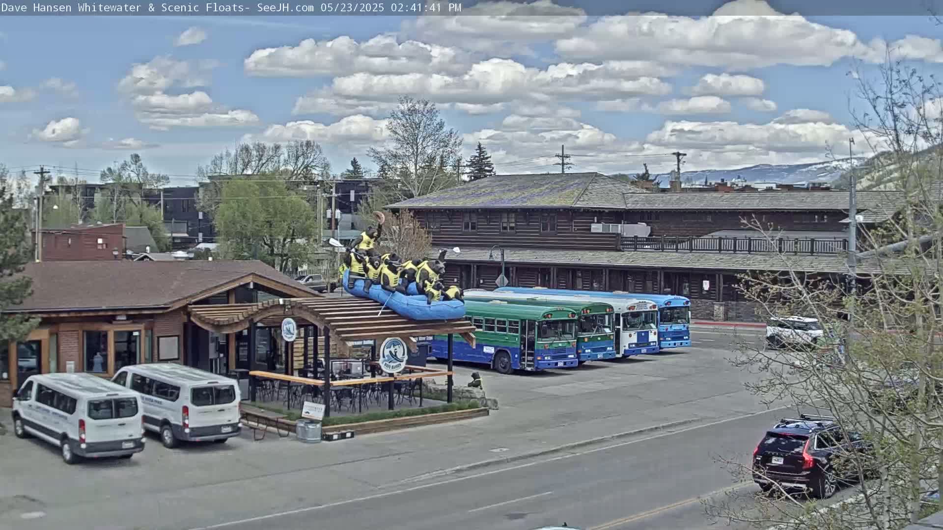 A partly cloudy day shows a building with a patio, two white vans, several buses parked nearby, and a raft with figures on top of the building's awning.