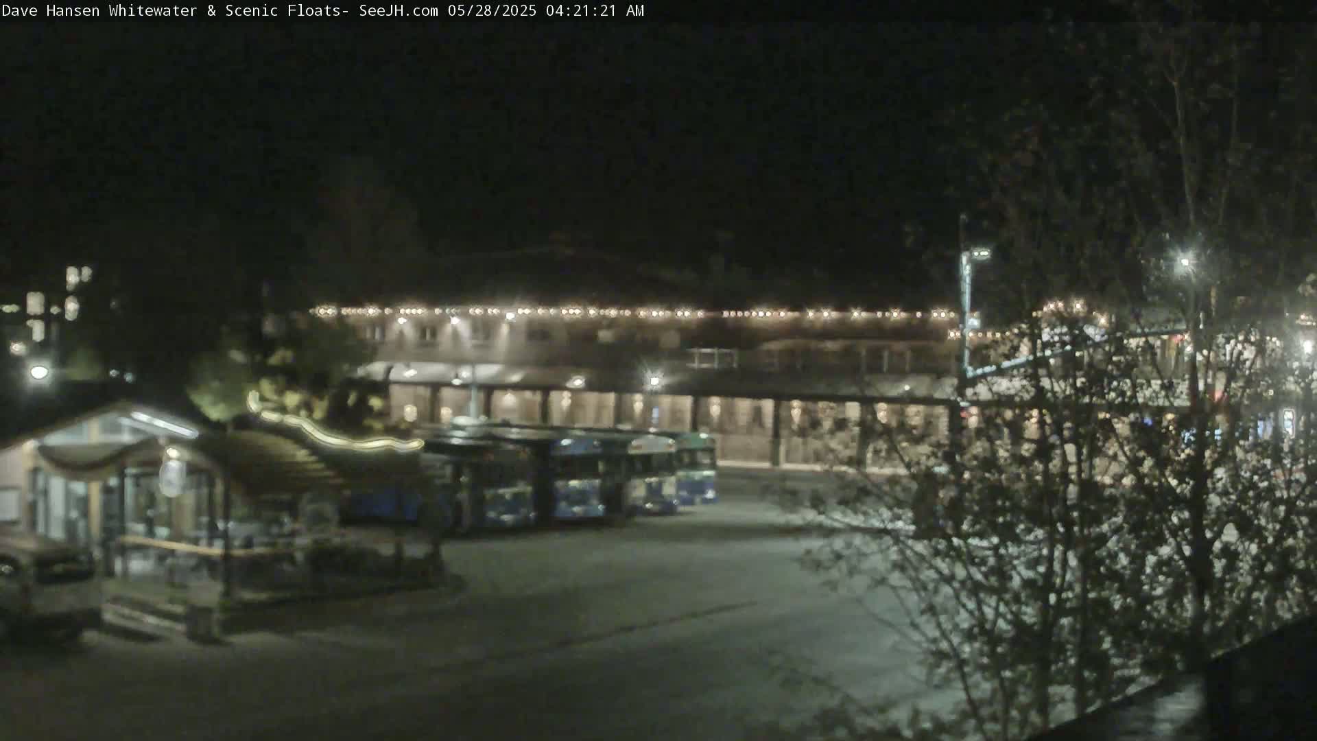 At night, several buses are parked in front of a building with lights on, and bare trees are visible in the foreground.