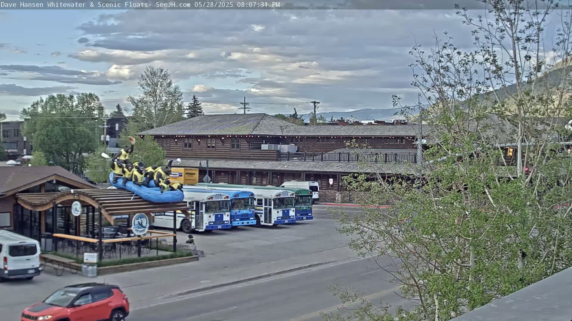 A partly cloudy afternoon scene shows a long, brown building with several buses parked in front, a raft with figures on top of a small building, and several vehicles parked along a street.
