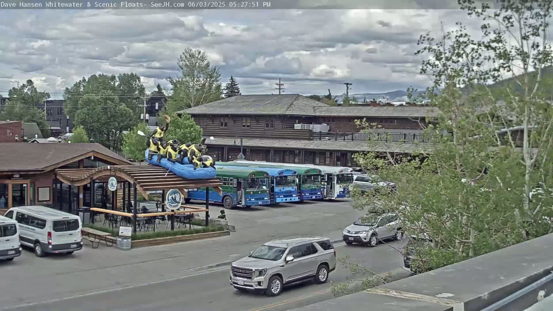 A partly cloudy day shows a group of people on a raft atop a building, several buses parked nearby, and various cars in a parking lot.
