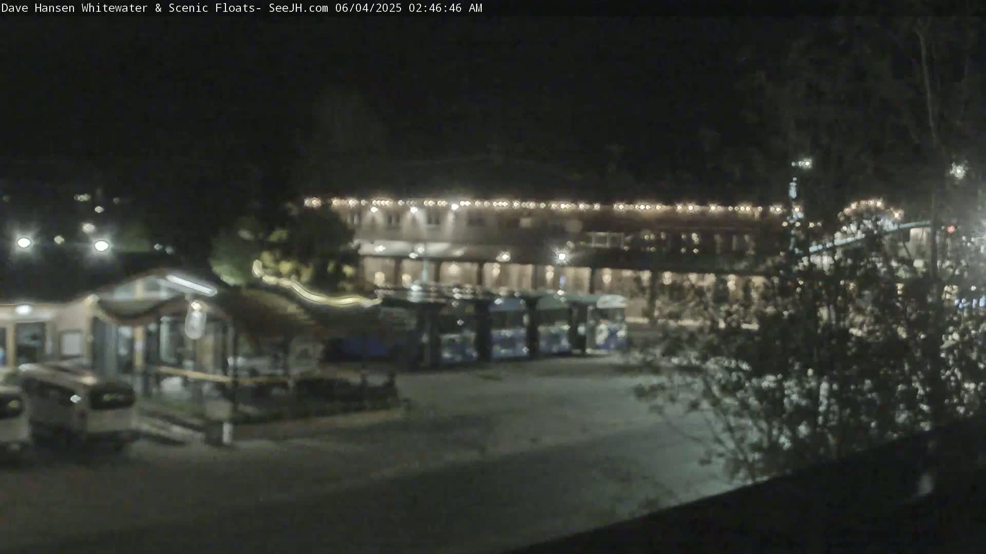 At night, several buses are parked in a lot next to a building with illuminated exterior lights.