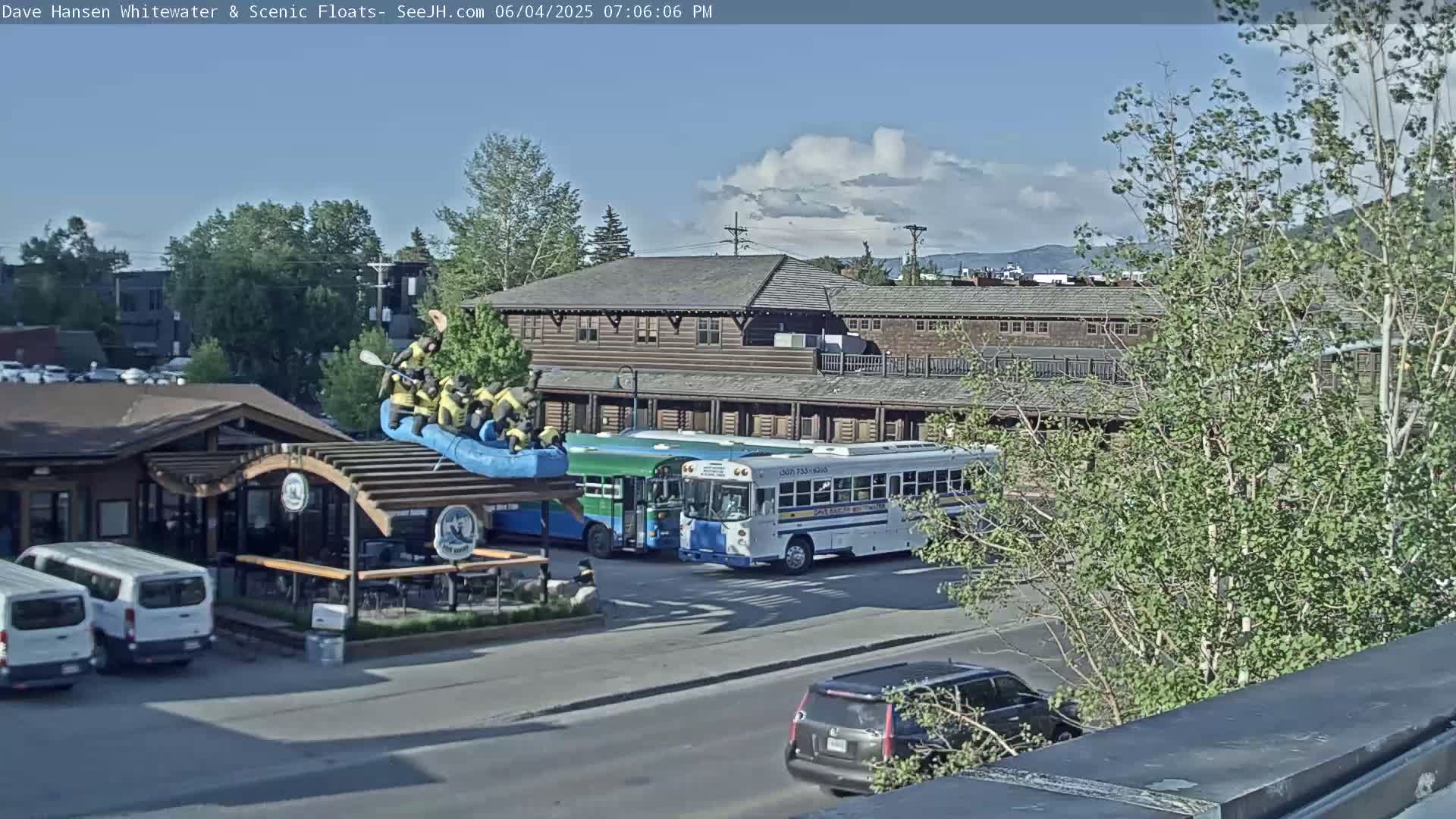 A partly cloudy day shows a view of a river rafting company with several people in a raft, parked buses, and cars in a parking lot near a long wooden building.