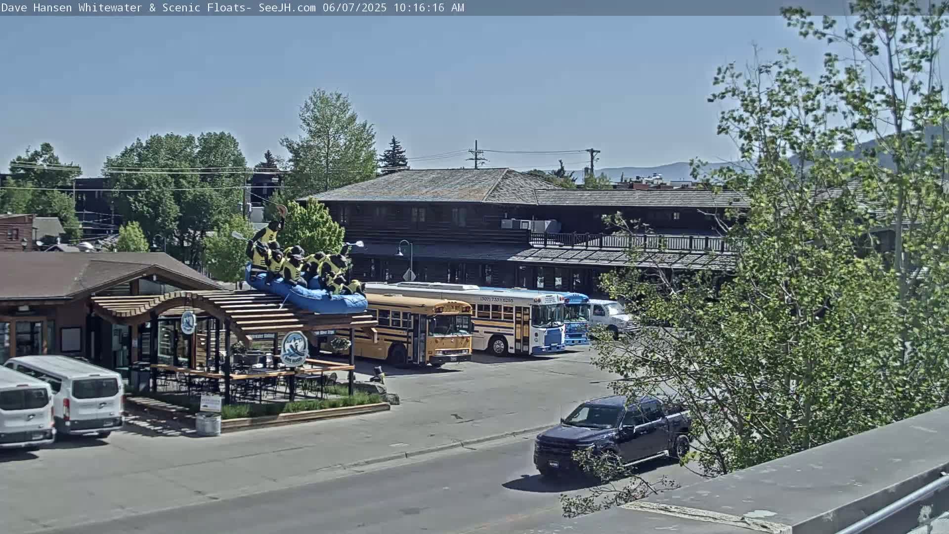 Under a clear sky, a parking lot contains several vehicles, including a school bus, near a building with a patio and a raft-shaped sculpture.