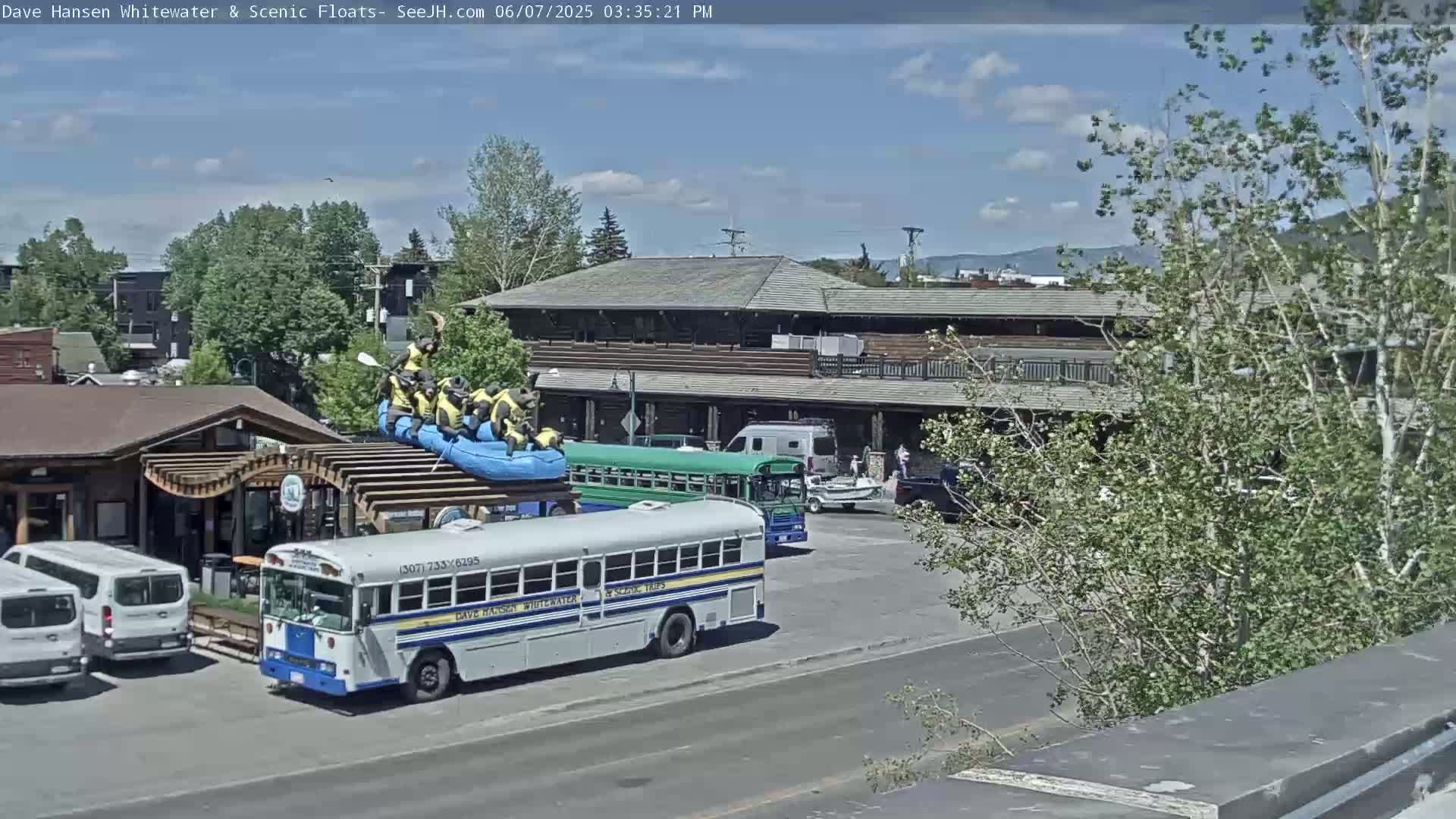 Under a partly sunny sky, a parking lot contains several vehicles, including two school buses and a raft with people on it, near a large building.