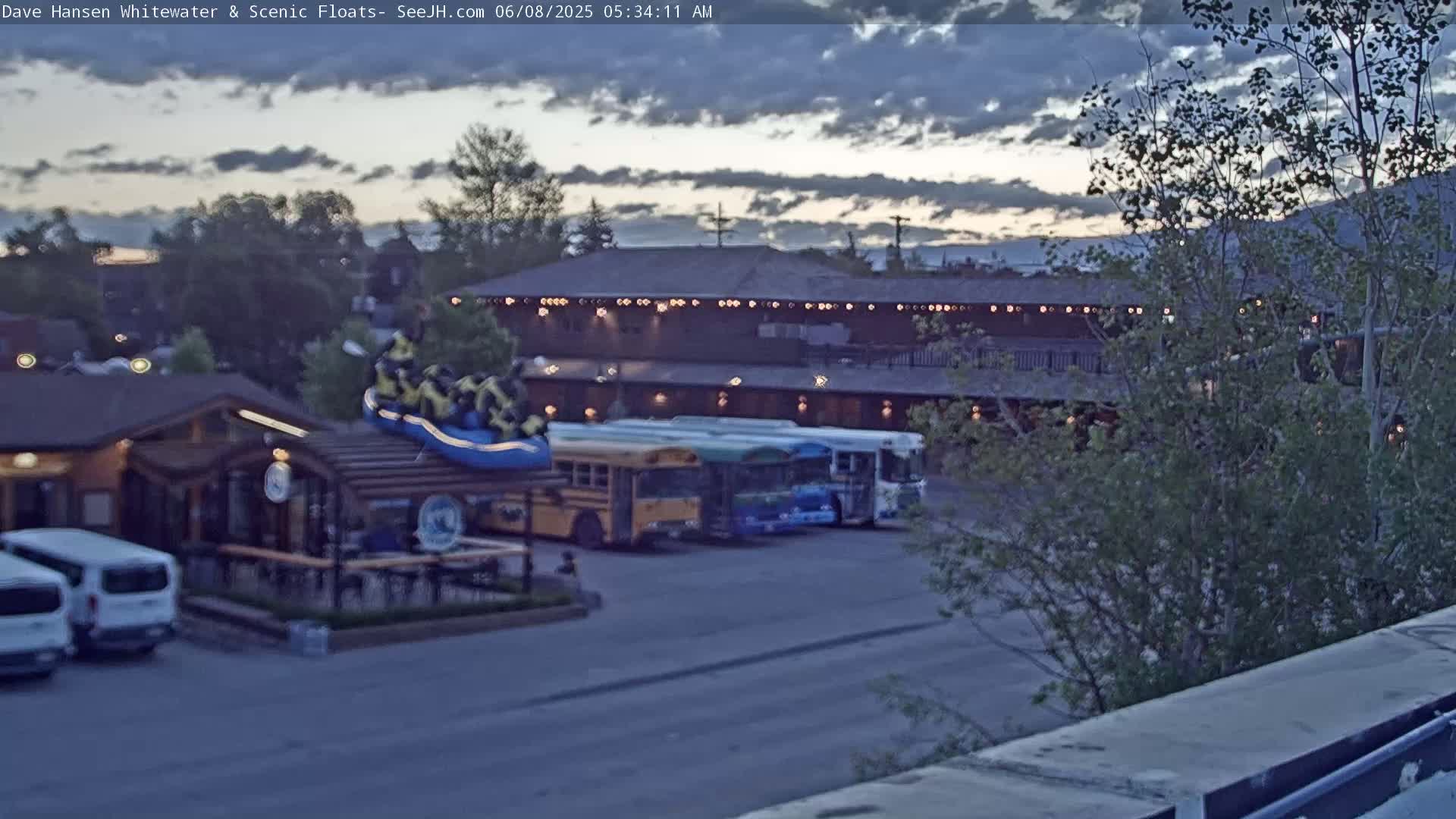 A group of people in a raft sits atop a building near several parked buses and vans under a partly cloudy twilight sky.