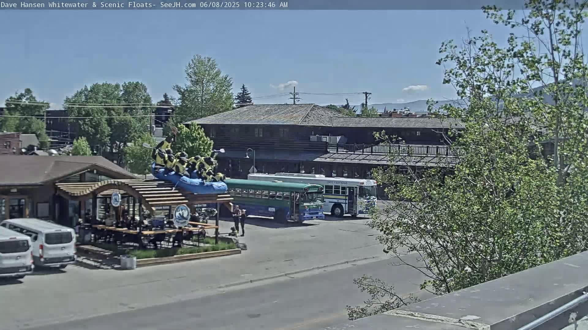 A sunny outdoor scene shows a group of people on a raft atop a building, a green school bus and a white bus parked in a lot, and several vans parked near a building with outdoor seating.