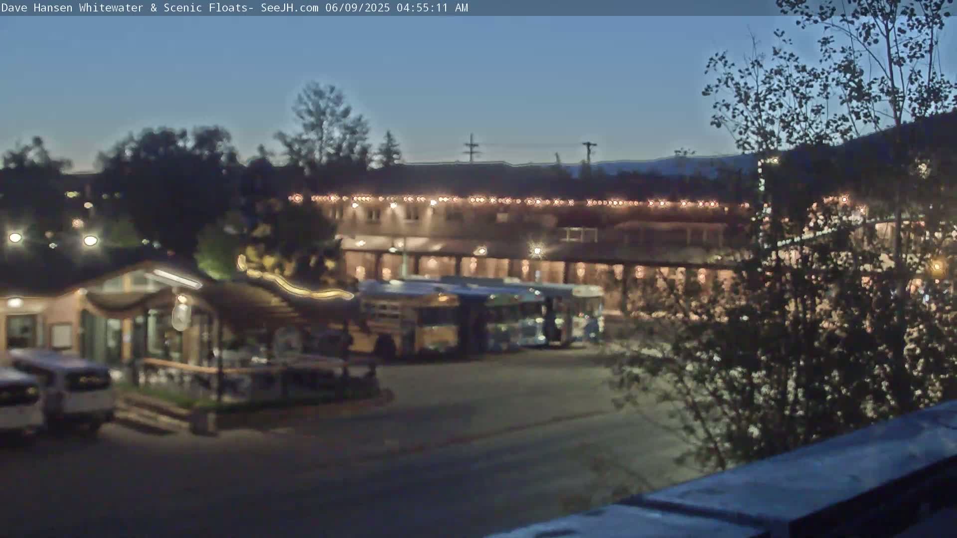A nighttime outdoor scene shows a building with lights, several buses parked in front, and trees in the foreground under a twilight sky.