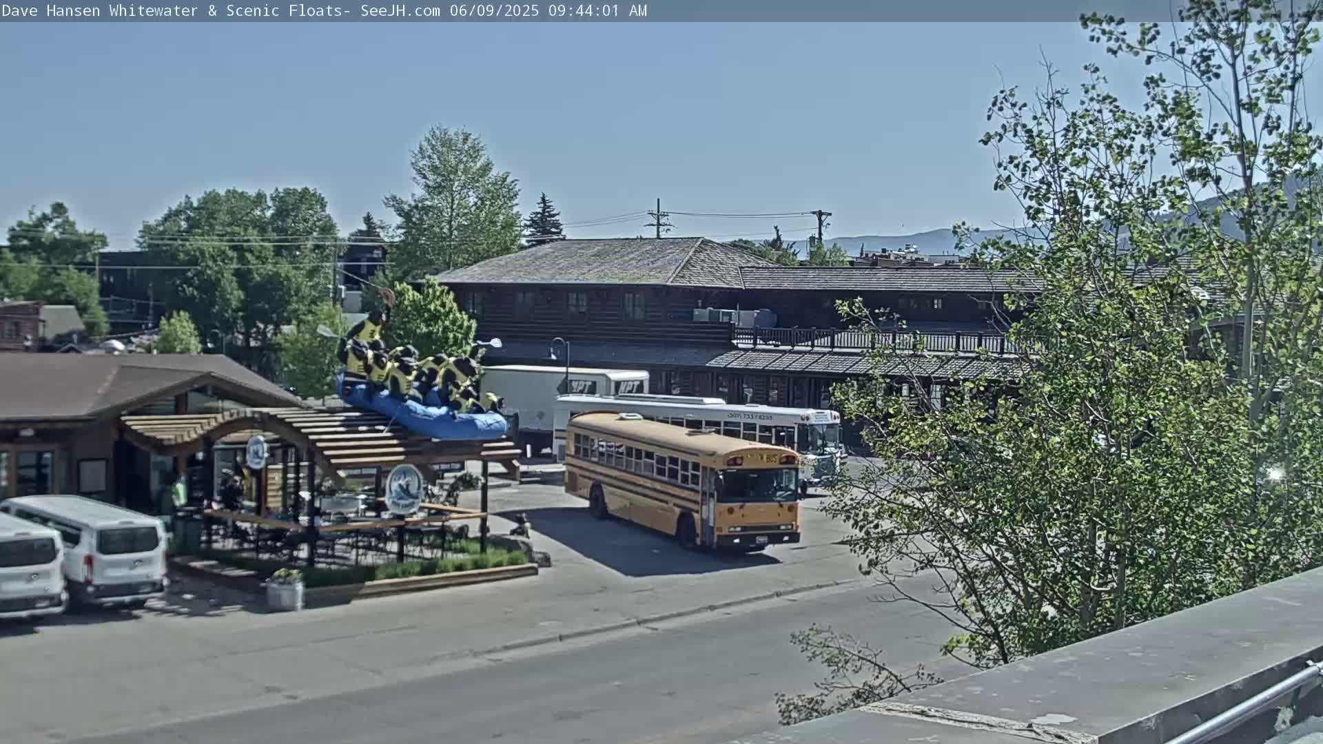 A sunny outdoor scene shows a yellow school bus parked near a building with a patio and a raft full of people on top of a structure.