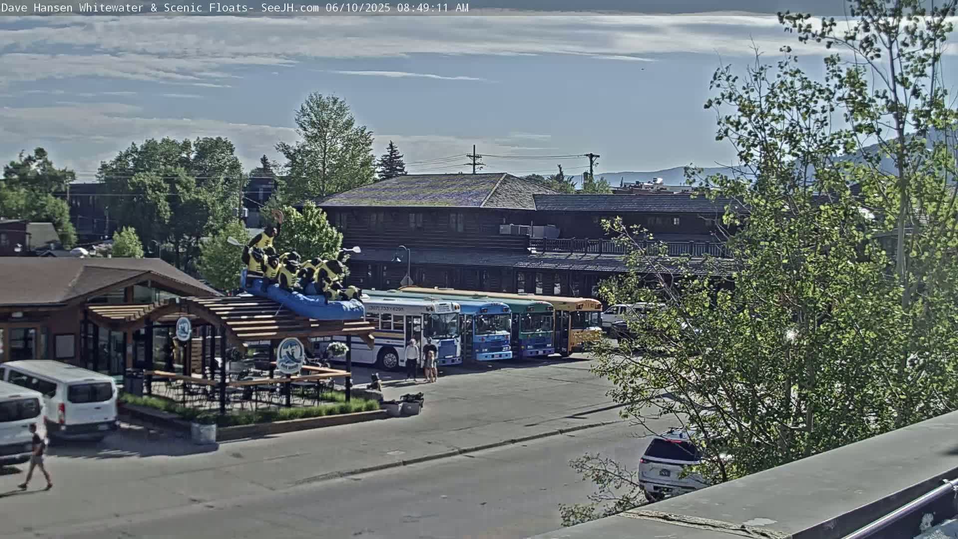 Under a partly cloudy sky, several colorful school buses are parked near a building with a large outdoor patio area and a whimsical inflatable raft display.