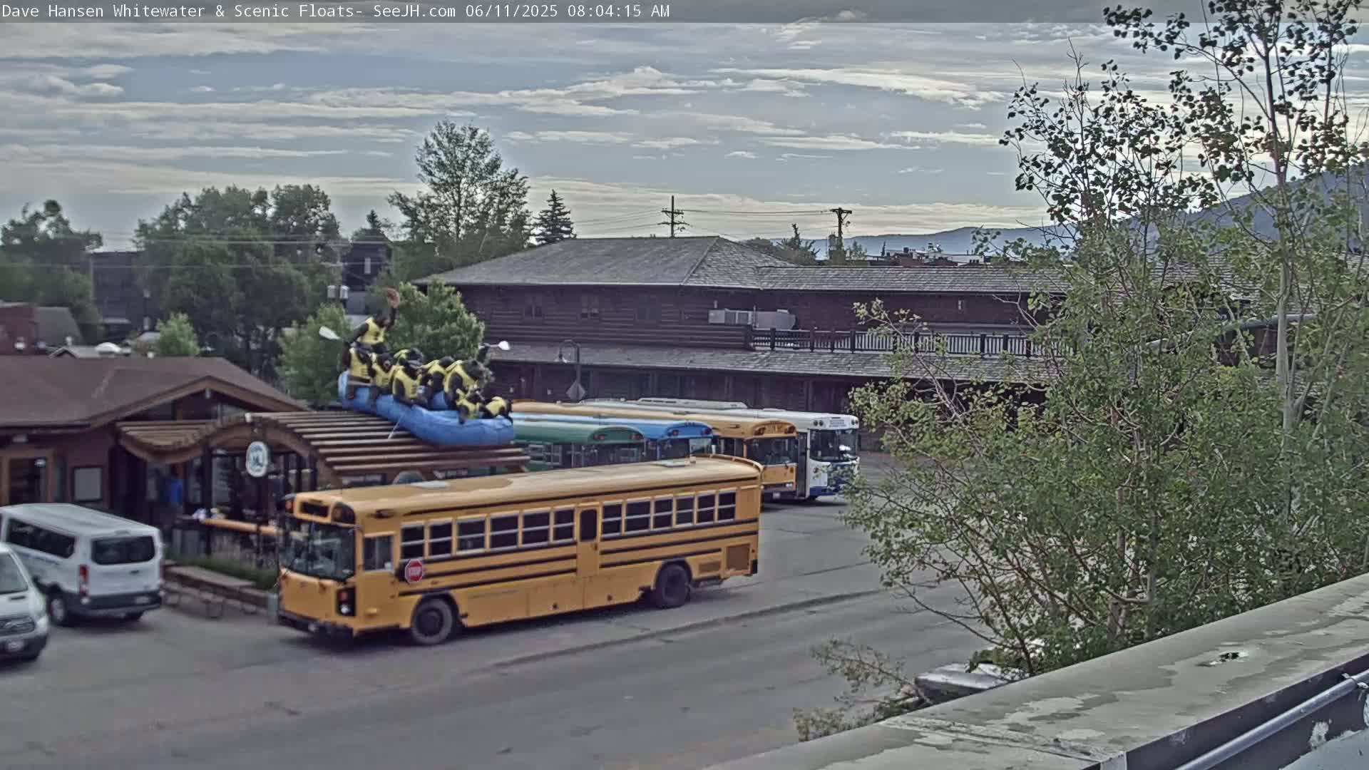 A partly cloudy morning scene shows a yellow school bus parked near several other buses and vans, with a group of people in a raft on a building's roof.