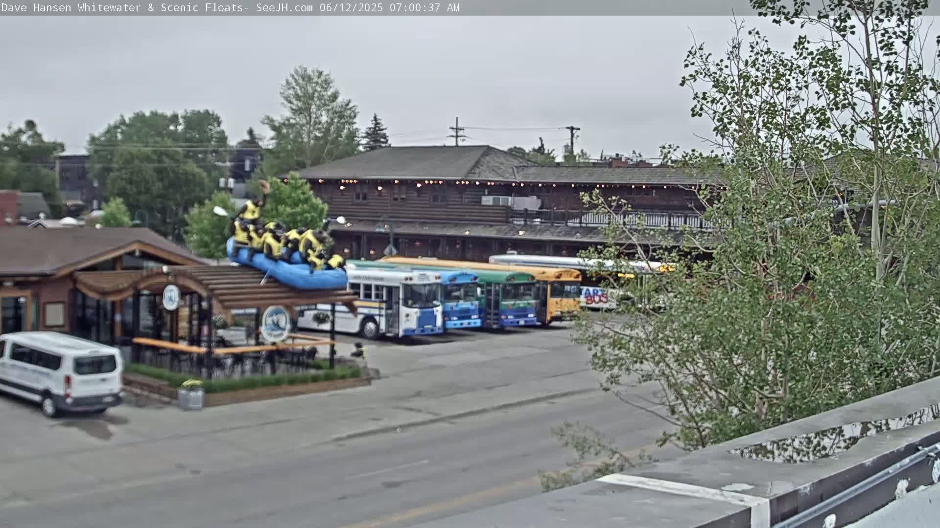 A group of people in yellow life vests are on a raft atop a building's roof, under an overcast sky, with several school buses parked nearby.