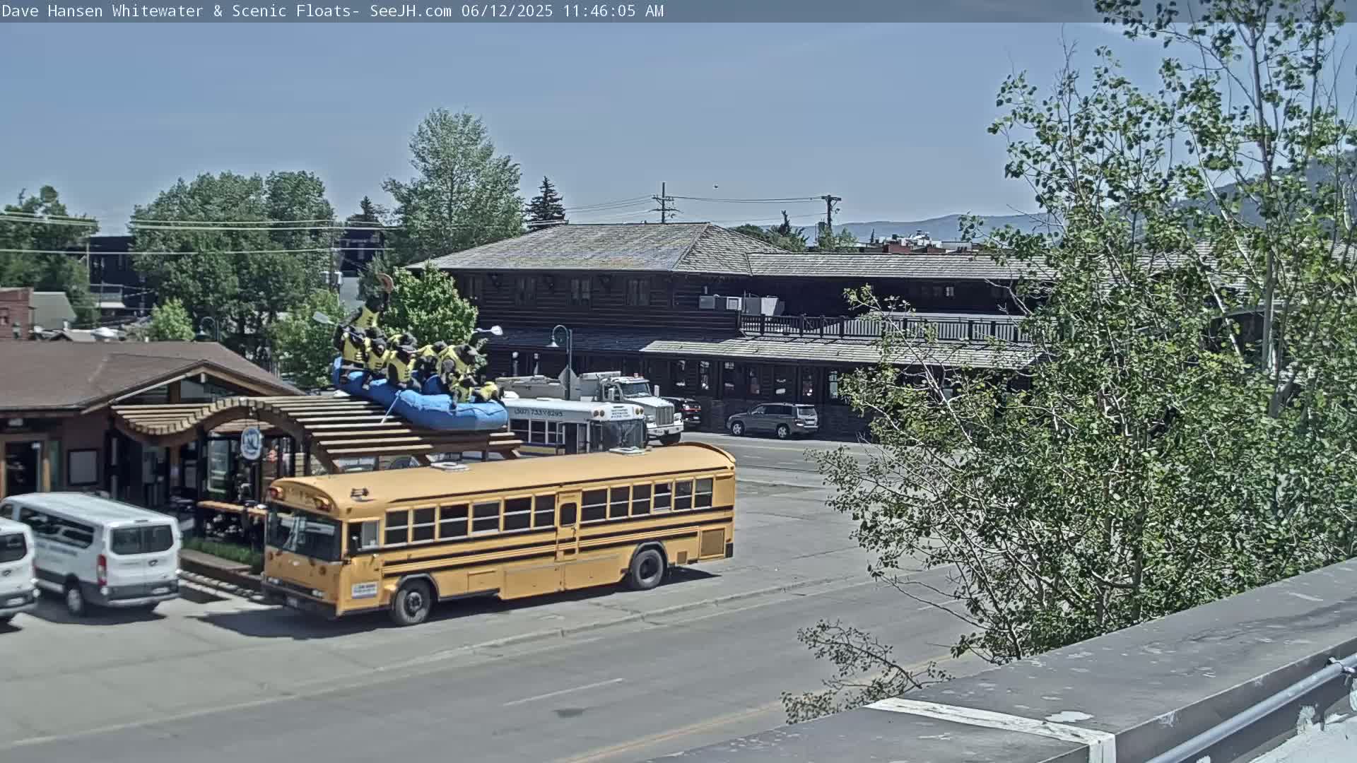 A yellow school bus with a raft full of people on its roof is parked in a sunny parking lot next to a building and some trees.