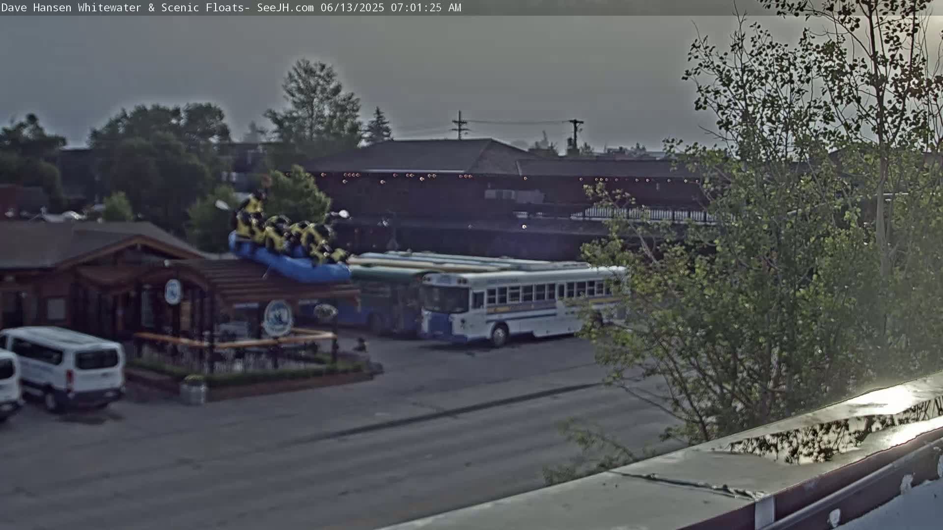 A hazy outdoor scene shows a group of people on a blue raft near a log cabin and several buses parked in a lot.