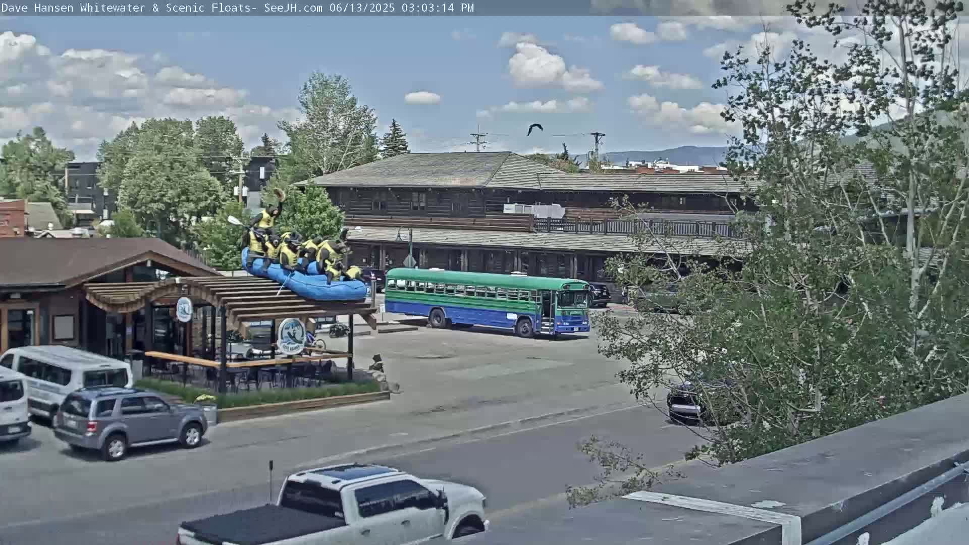 A partly cloudy day shows a group of people on a raft atop a structure near a parking lot with cars and a green and blue bus.