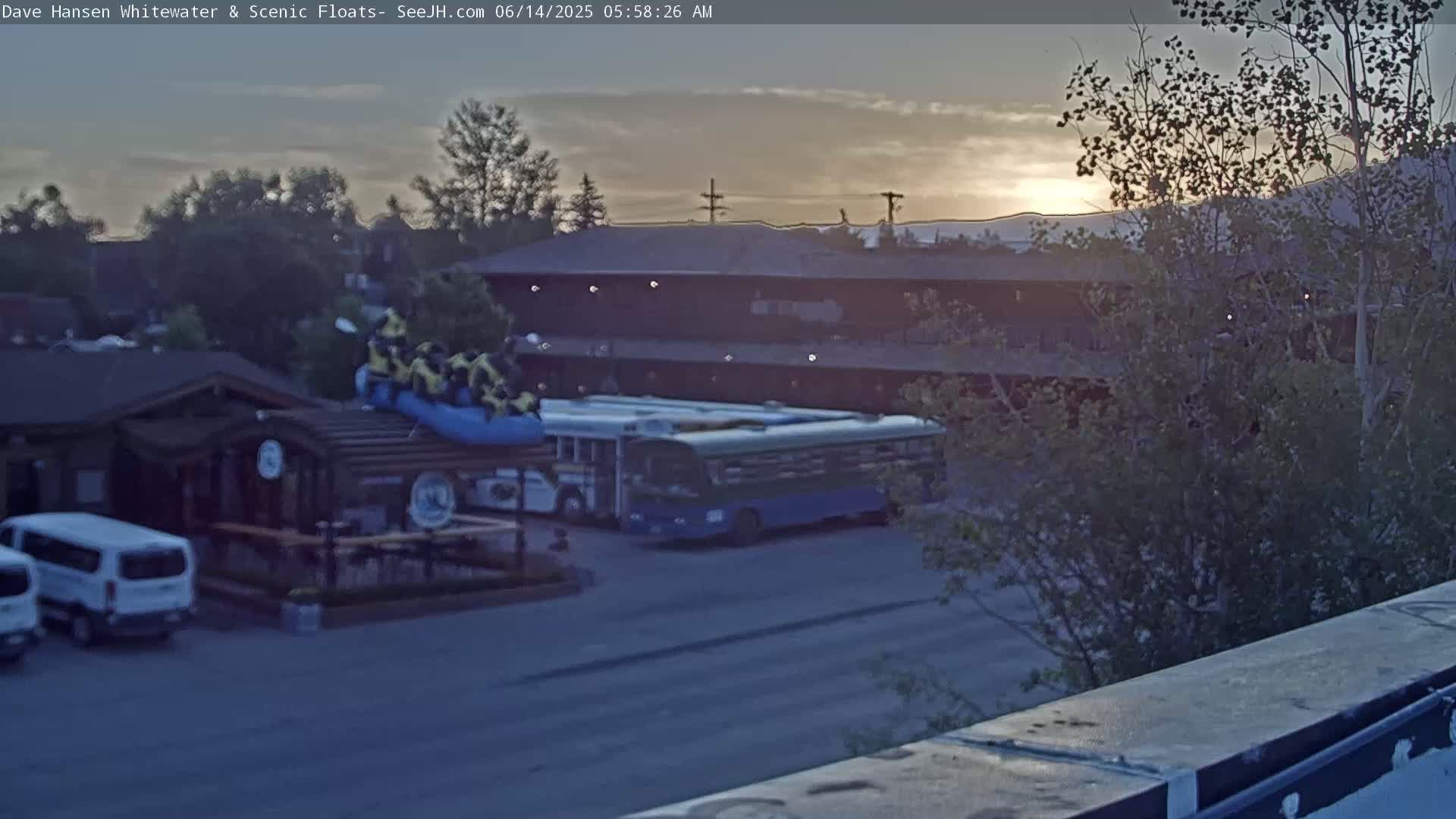 A sunrise illuminates a parking lot with several vehicles, including a bus, near a building and a raft with people on top of a building.