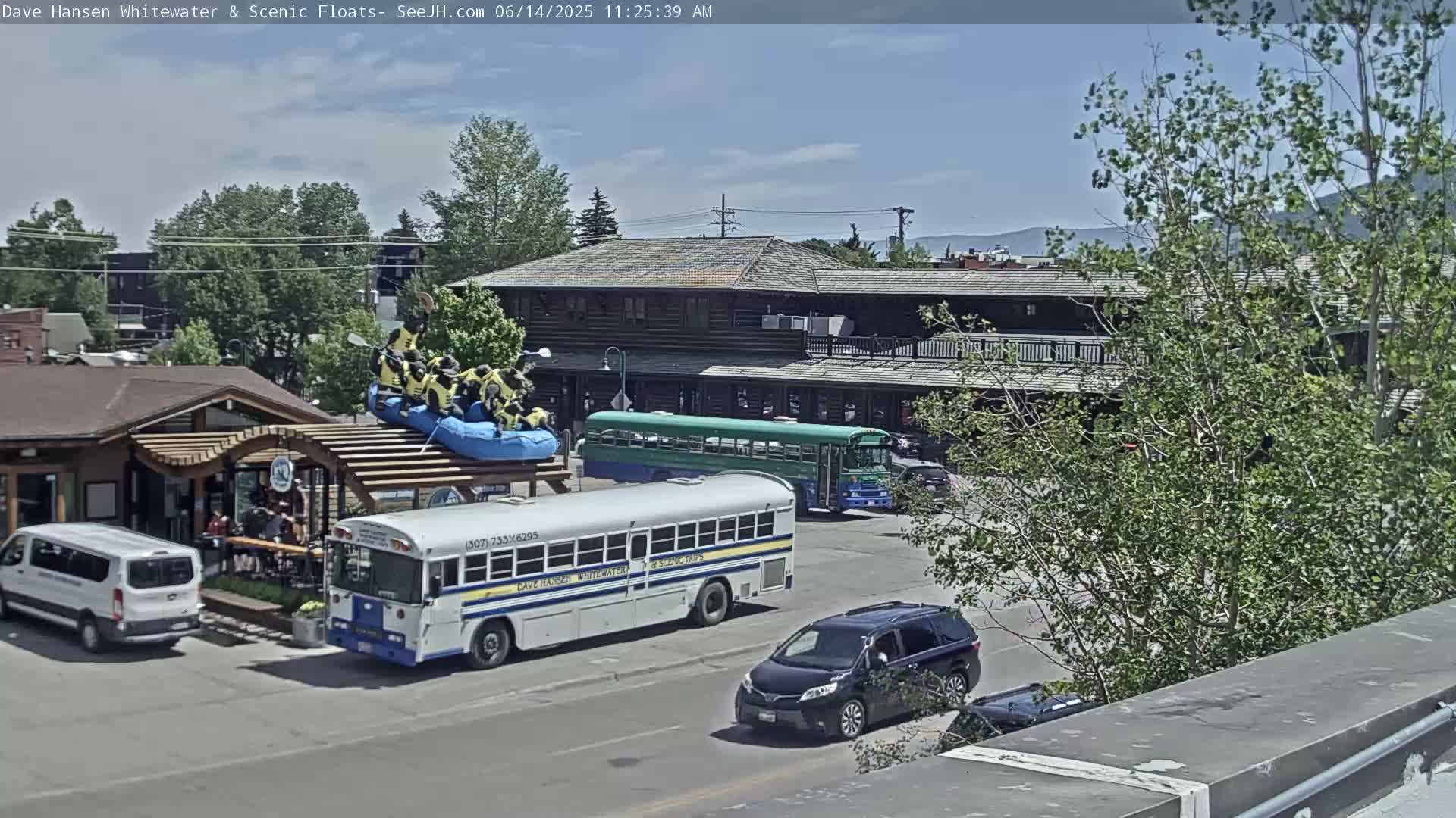 A sunny day shows a parking lot with several vehicles, including two buses and a raft with people in life vests on top of a building, near a wood-framed building with a second story.