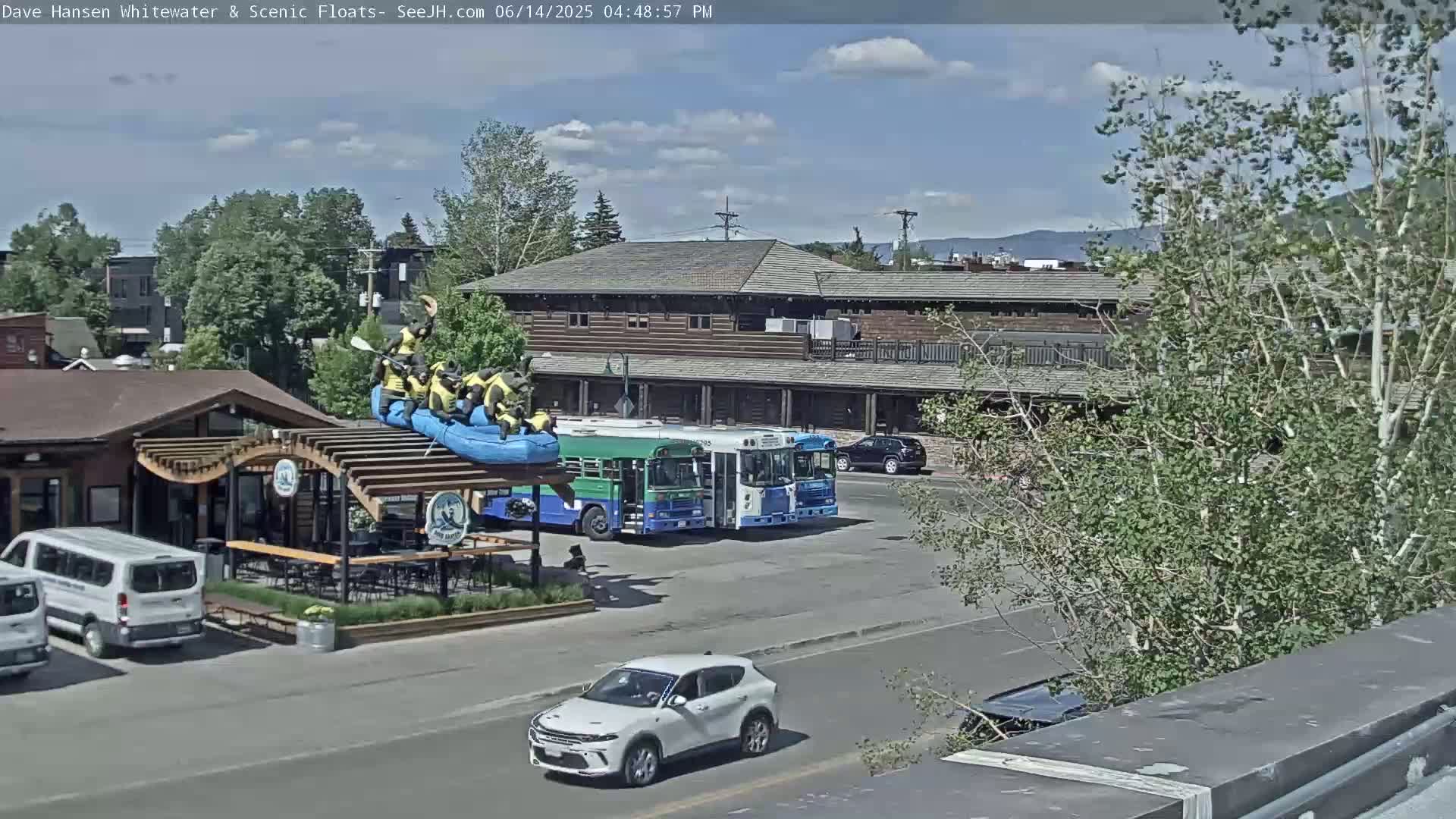 A sunny day shows a parking lot with several vehicles, including two buses and a raft with people in life vests on top of a building, near a wood-framed building with a second story.