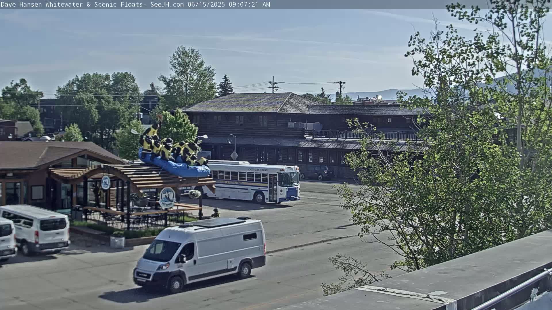 A sunny day reveals a parking lot with a white van, a school bus, several white vans, and a log building with a rafting inflatable on its roof, all surrounded by lush green trees and a mountain range in the distance.