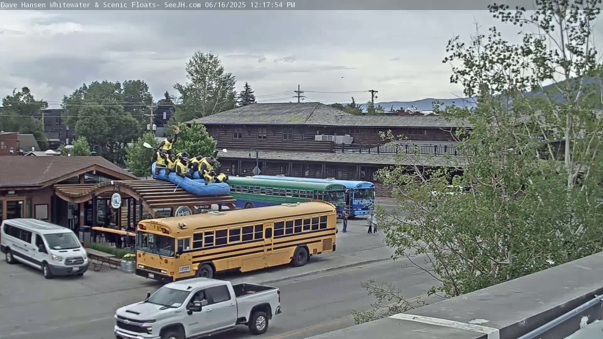 A group of people in yellow life vests are sitting on a raft on top of a building, near two school buses and other vehicles, under an overcast sky.