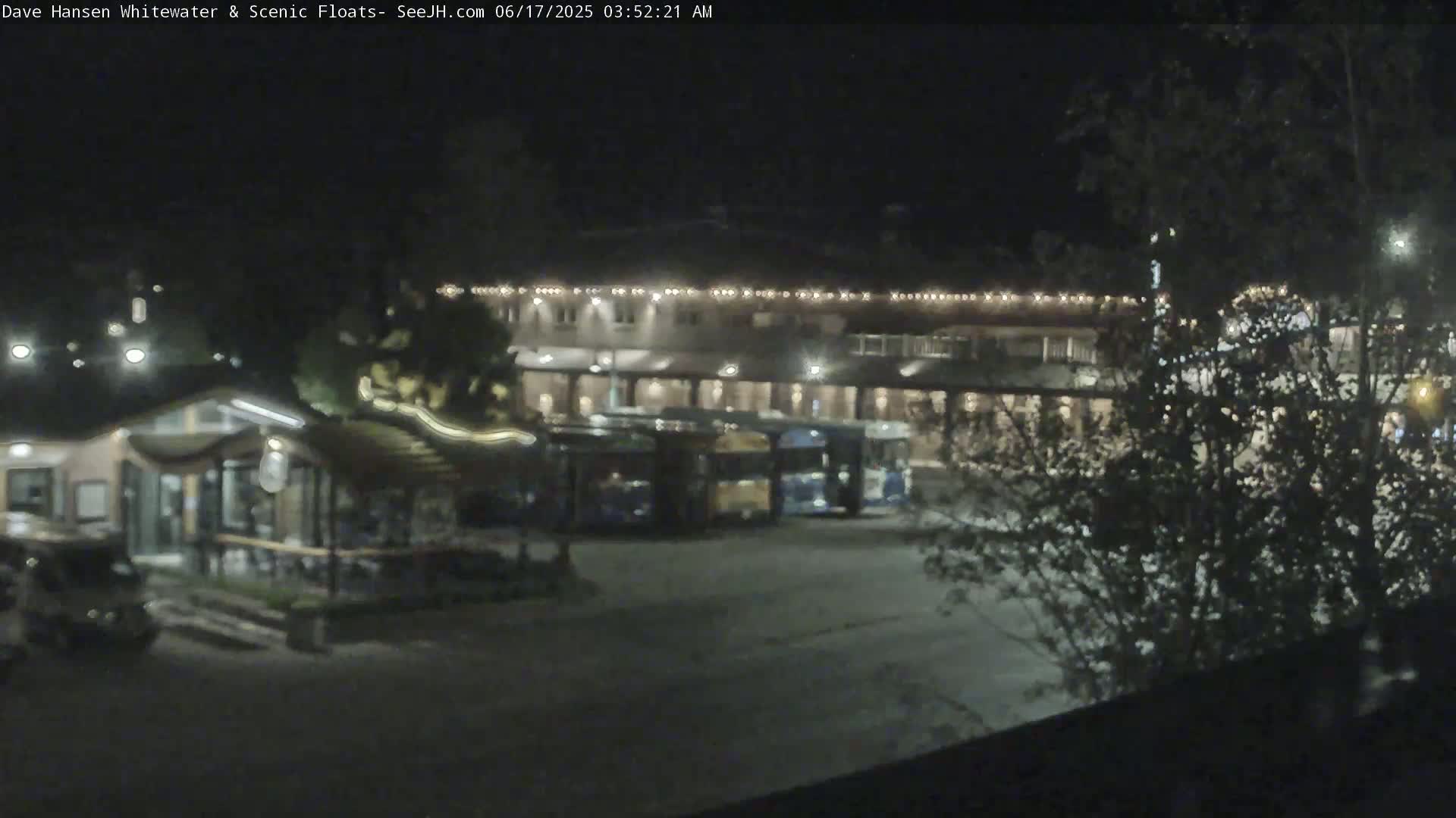 At night, several buses are parked in a lot next to a building with lights on, surrounded by trees.