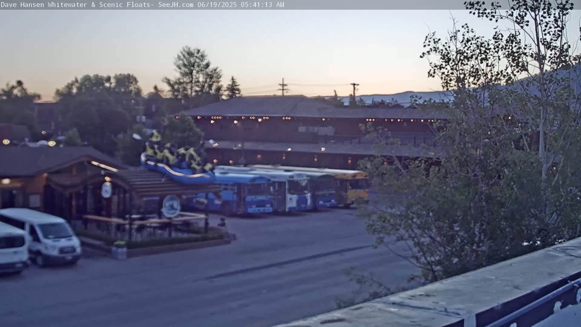 A twilight view shows a parking lot with several buses, a log cabin-style building with a patio, and a large inflatable raft near the building.