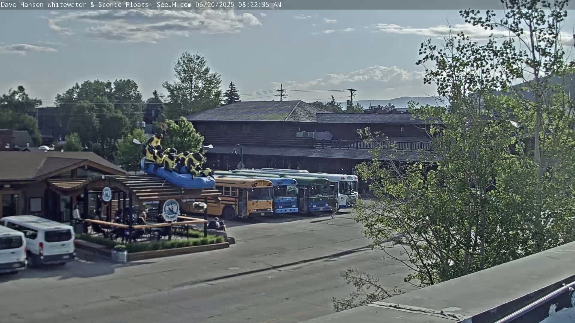 A partly cloudy day shows a group of people on a raft atop a building near several buses and vans parked in a lot next to a log cabin-style building.