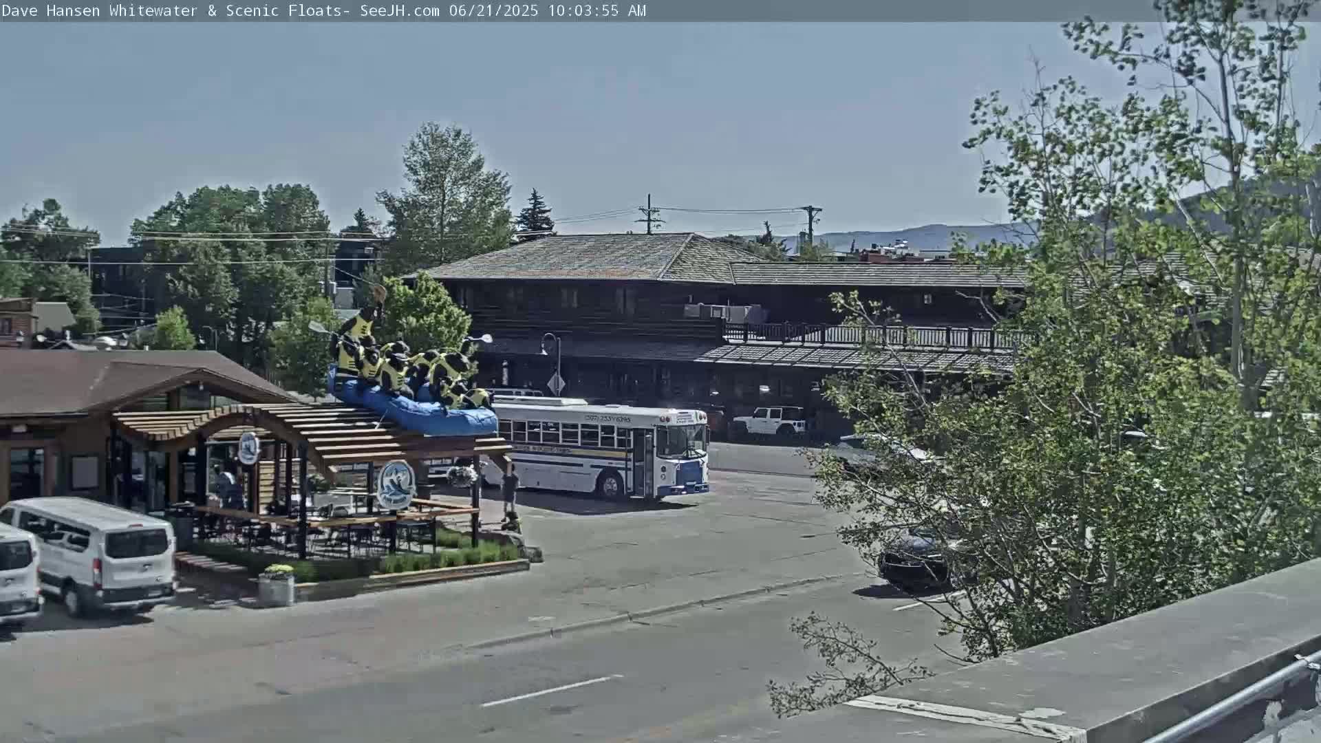 A sunny day reveals a street scene with a large building, a school bus, several vehicles, and a raft with people on top situated near a restaurant with outdoor seating.