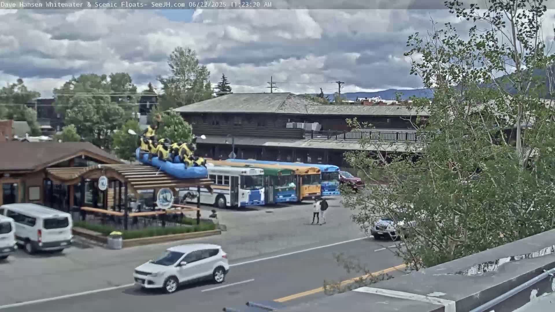 A group of people in yellow life vests sits on a blue raft atop a building, under a partly cloudy sky, near several parked buses and cars.