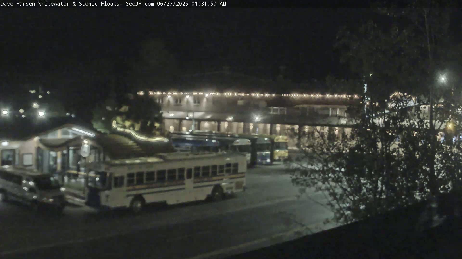 At night, several buses are parked in a lot next to a building with lights outlining its roofline, partially obscured by trees.