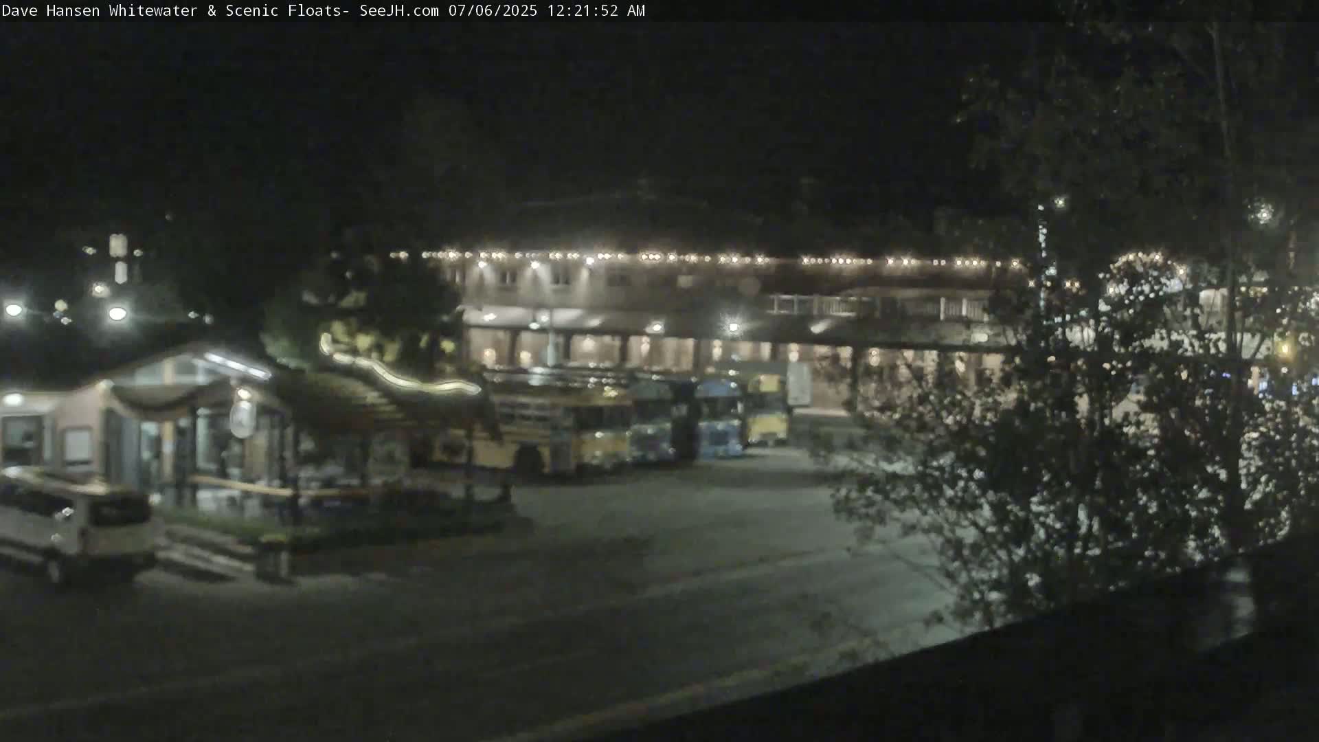 At night, a parking lot with several buses and a building illuminated with string lights is partially obscured by trees.