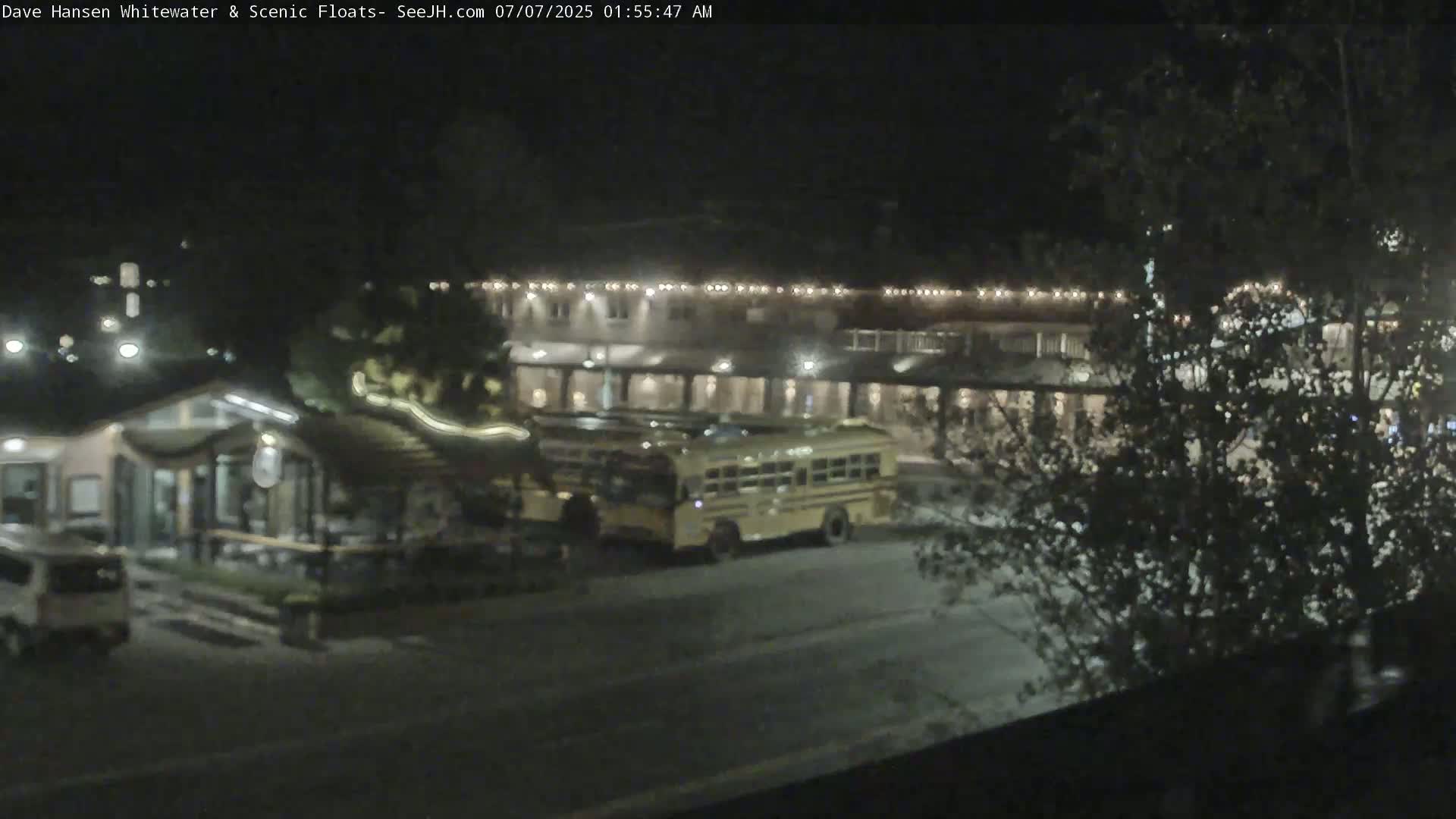 At night, two school buses are parked in front of a brightly lit building next to a smaller structure, all surrounded by dark trees.