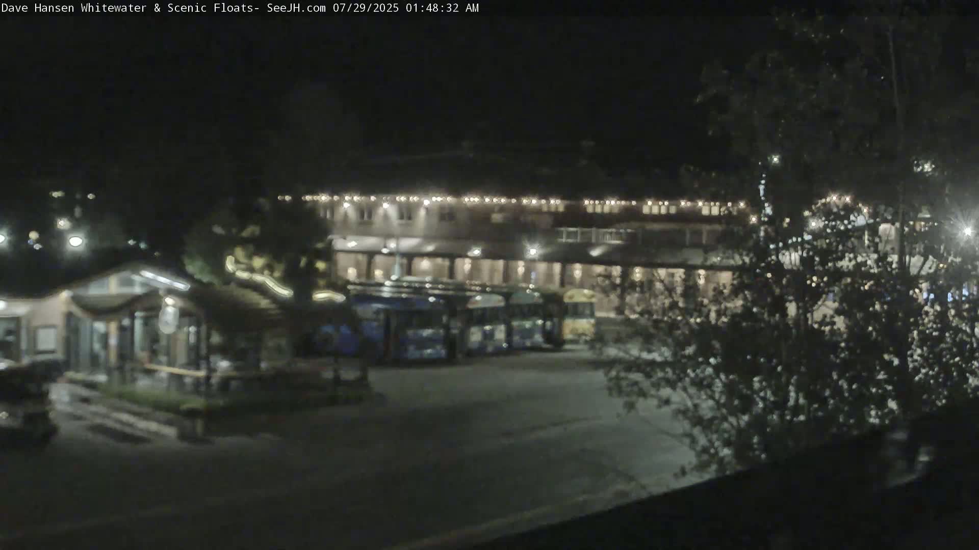A nighttime view shows a parking lot with several buses parked in front of a large building illuminated by lights, surrounded by trees.