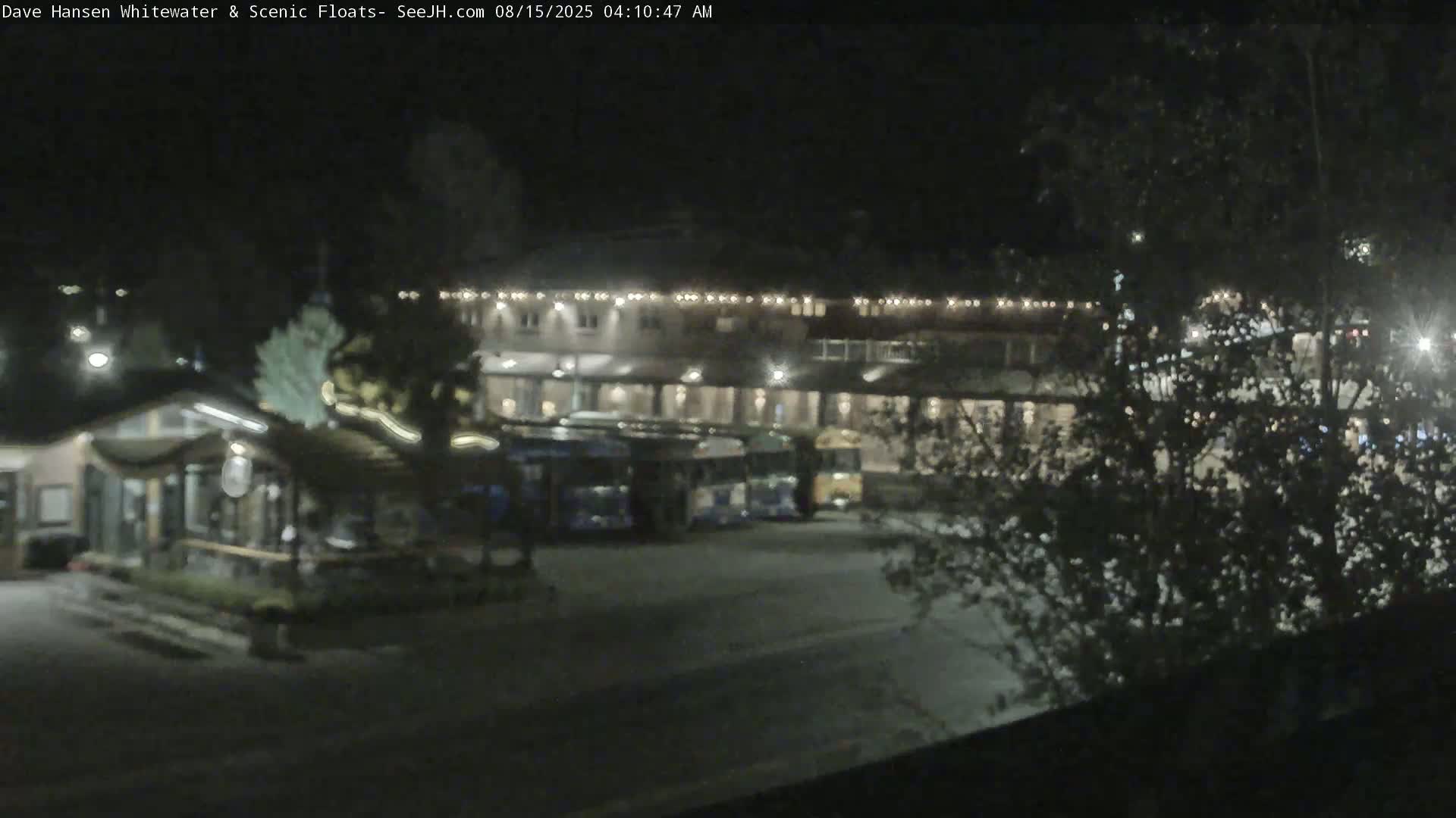 A nighttime view shows a parking lot with several buses parked in front of a large building illuminated by lights, surrounded by trees.