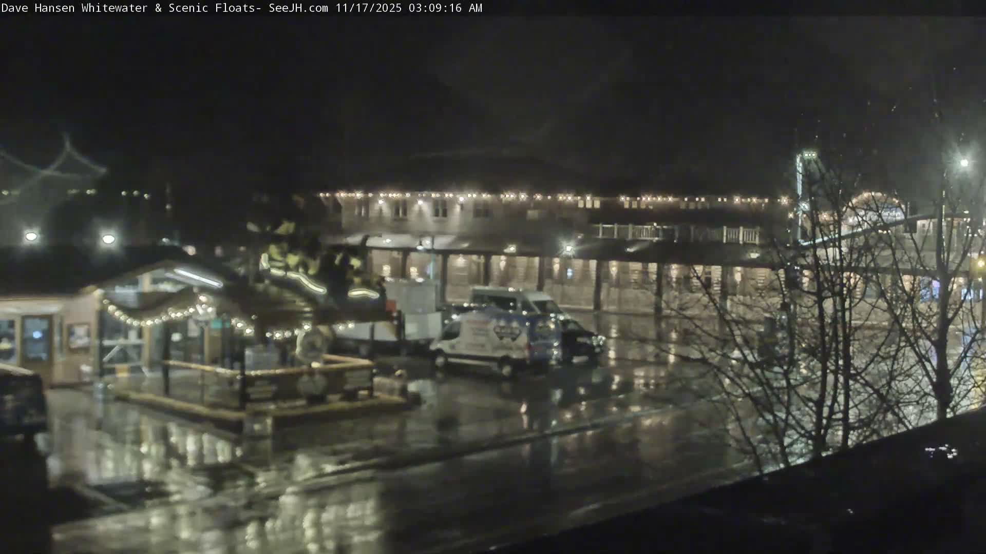 A nighttime scene depicts a wet town street with vehicles, a small building adorned with string lights, and a multi-story illuminated building in the background, all under rainy conditions.