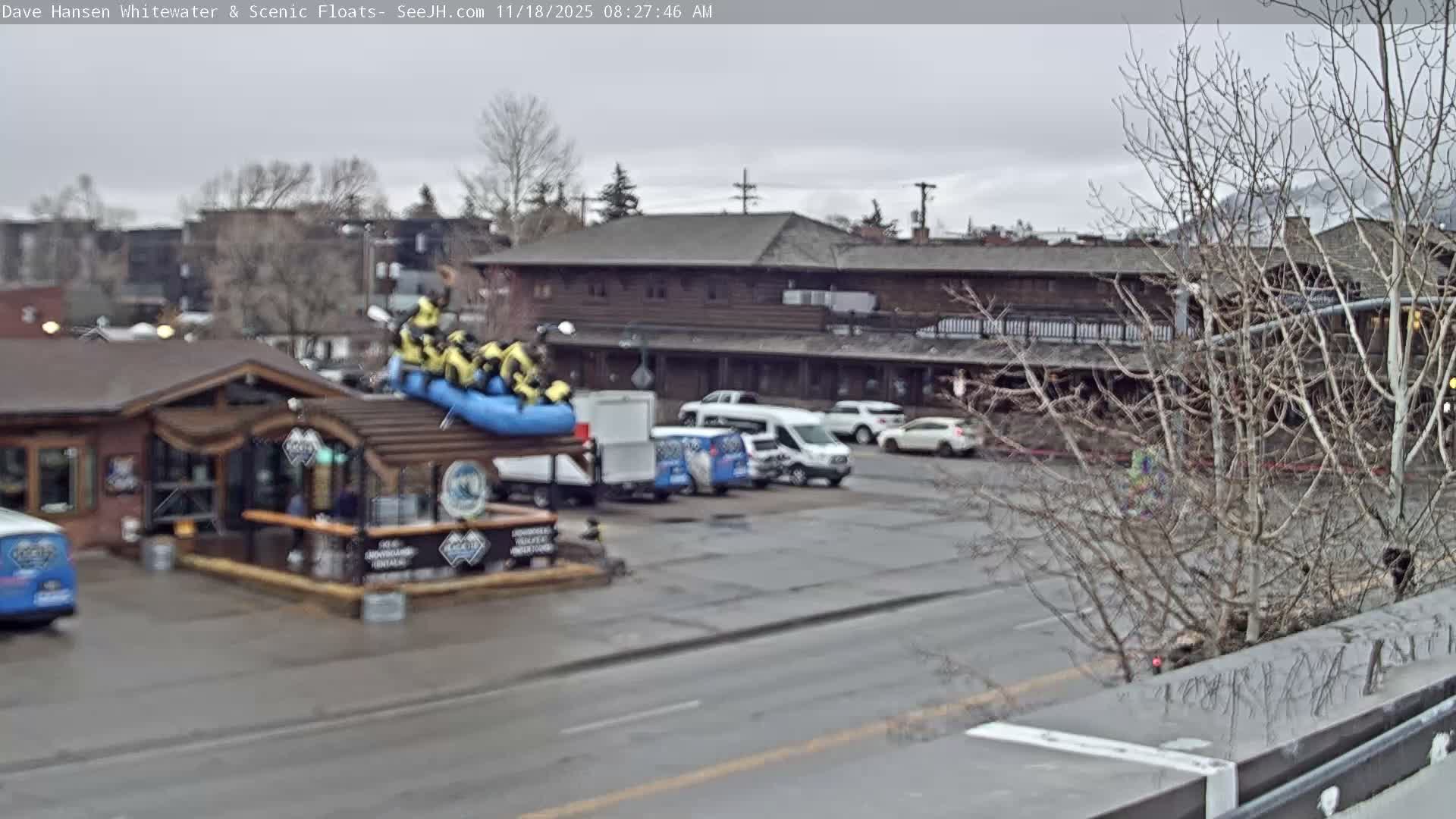 A nighttime scene depicts a wet town street with vehicles, a small building adorned with string lights, and a multi-story illuminated building in the background, all under rainy conditions.